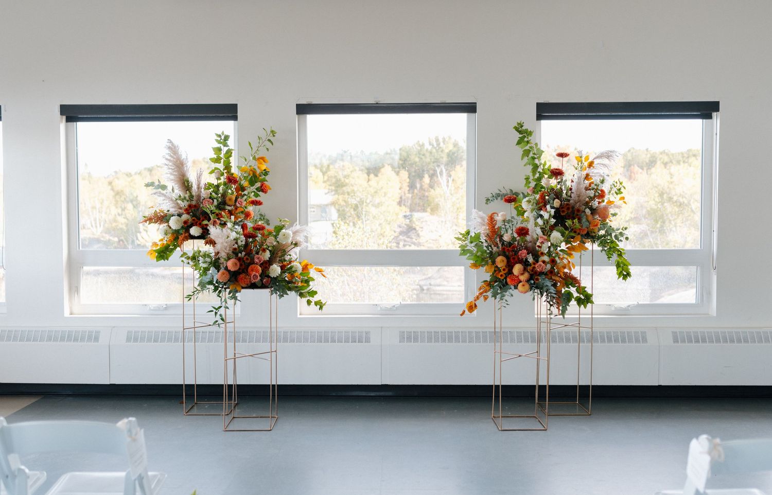 Four tall gold stand floral arrangements with orange, white, and green flowers displayed in a bright white ceremony venue.