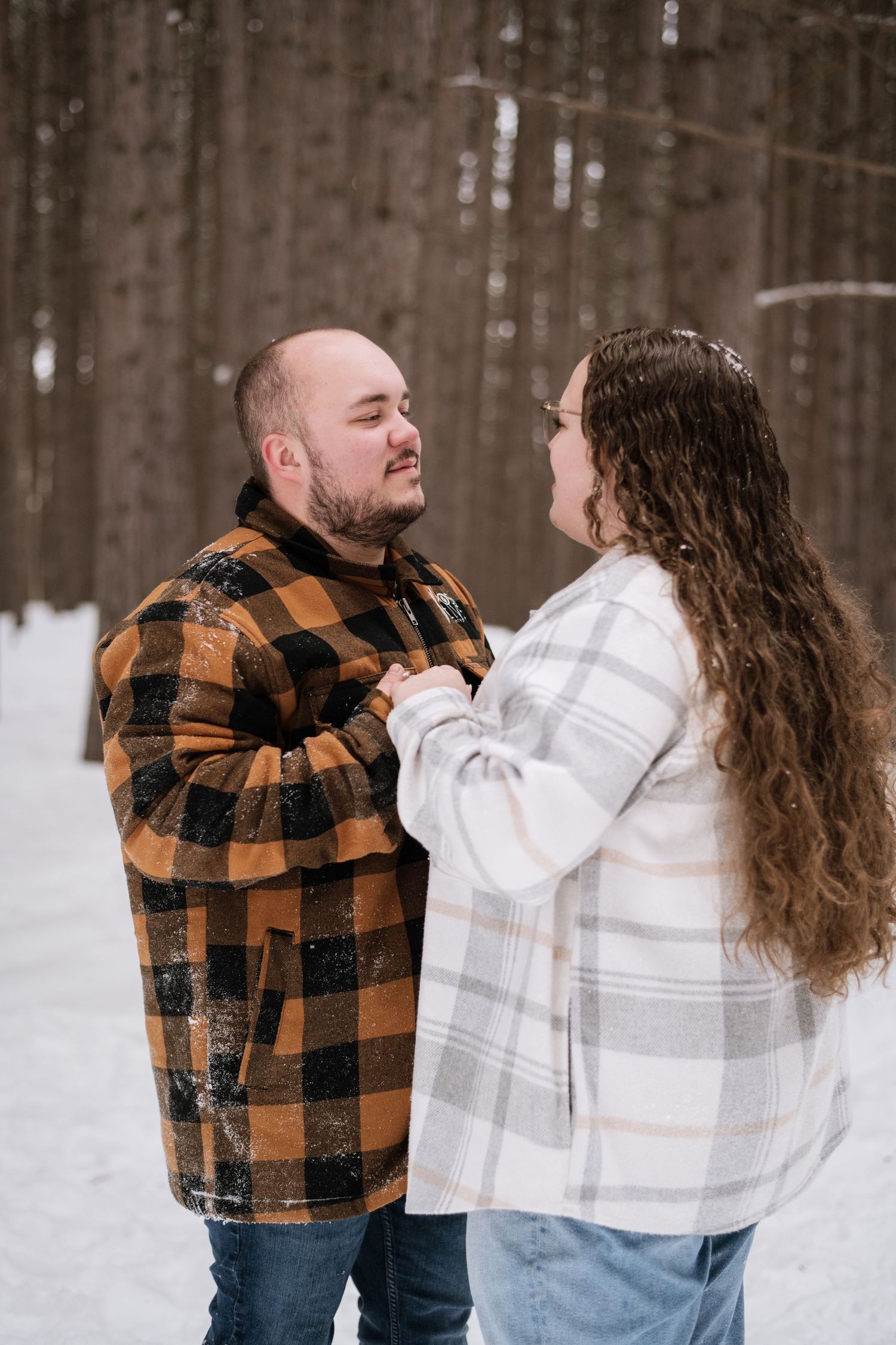 Couple in plaid jackets sharing a tender moment in a snowy winter forest setting.