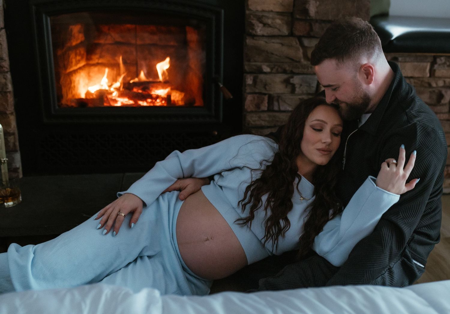 Couple cuddling on bed in front of fireplace during intimate maternity photoshoot.