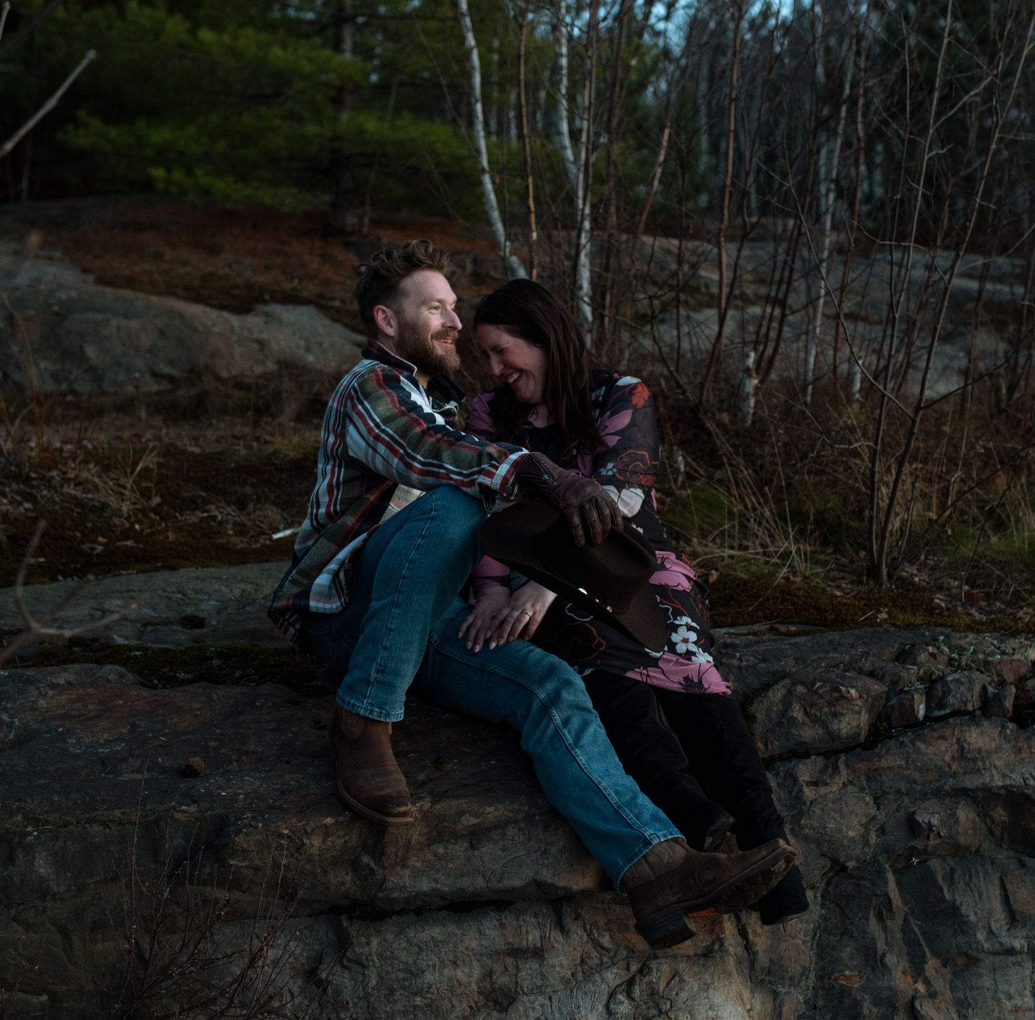 A couple sits together on a rocky ledge at dusk surrounded by birch trees in a moody forest setting.