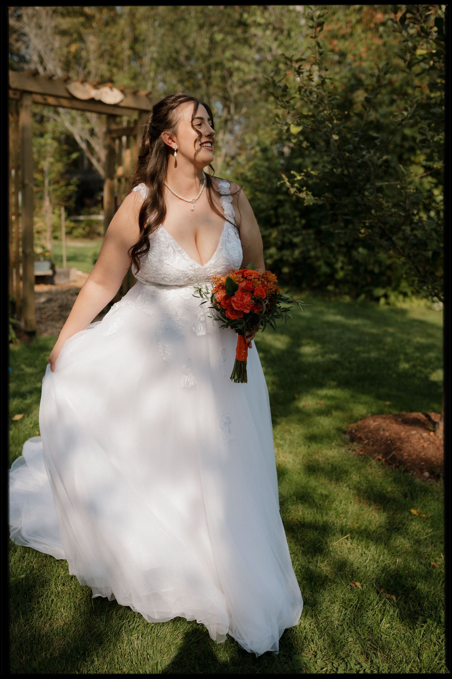 A bride in a flowing white gown holds a vibrant red bouquet while standing in a sunlit garden setting.