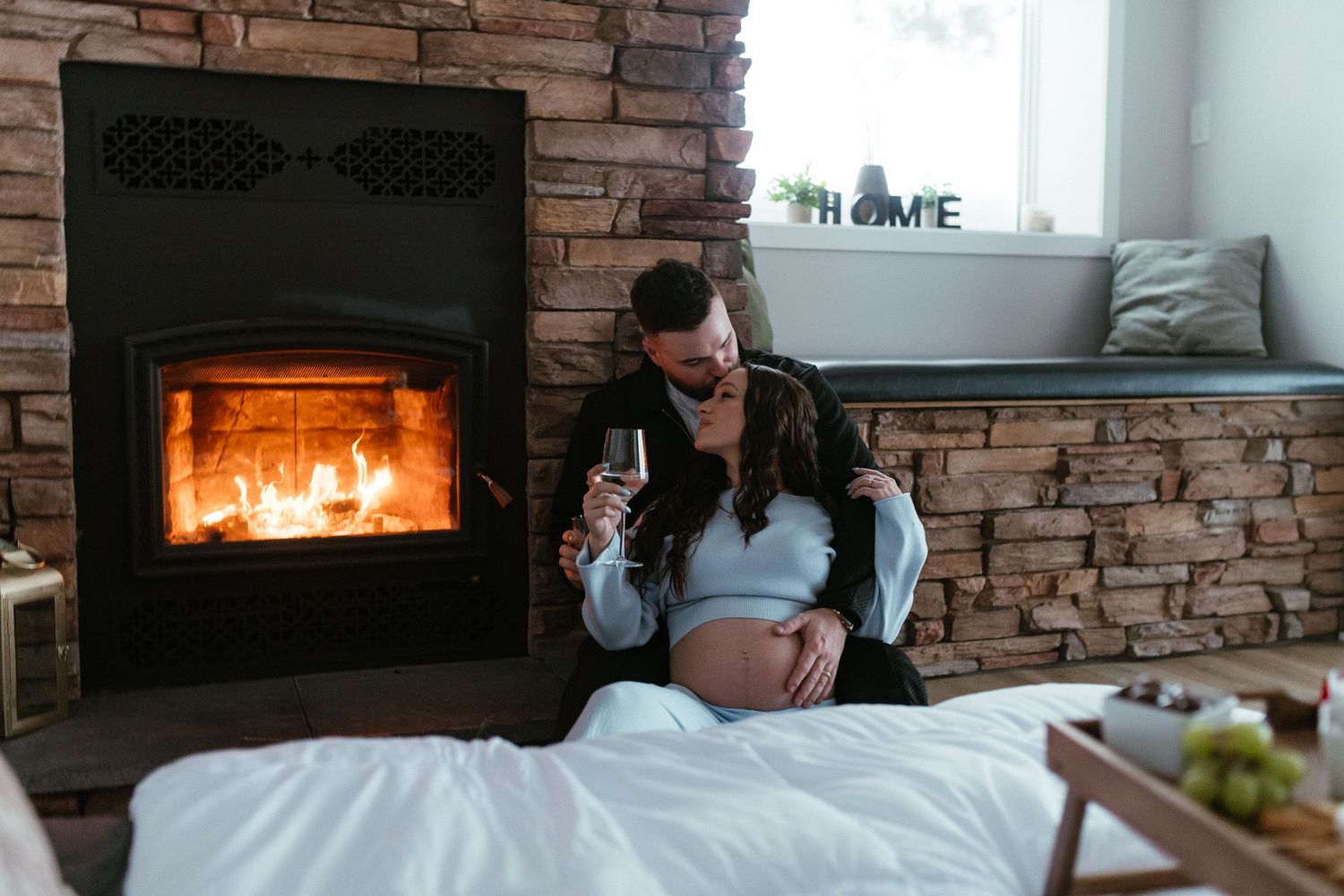 Couple relaxing by cozy fireplace, man embracing pregnant woman on bed in rustic living room.