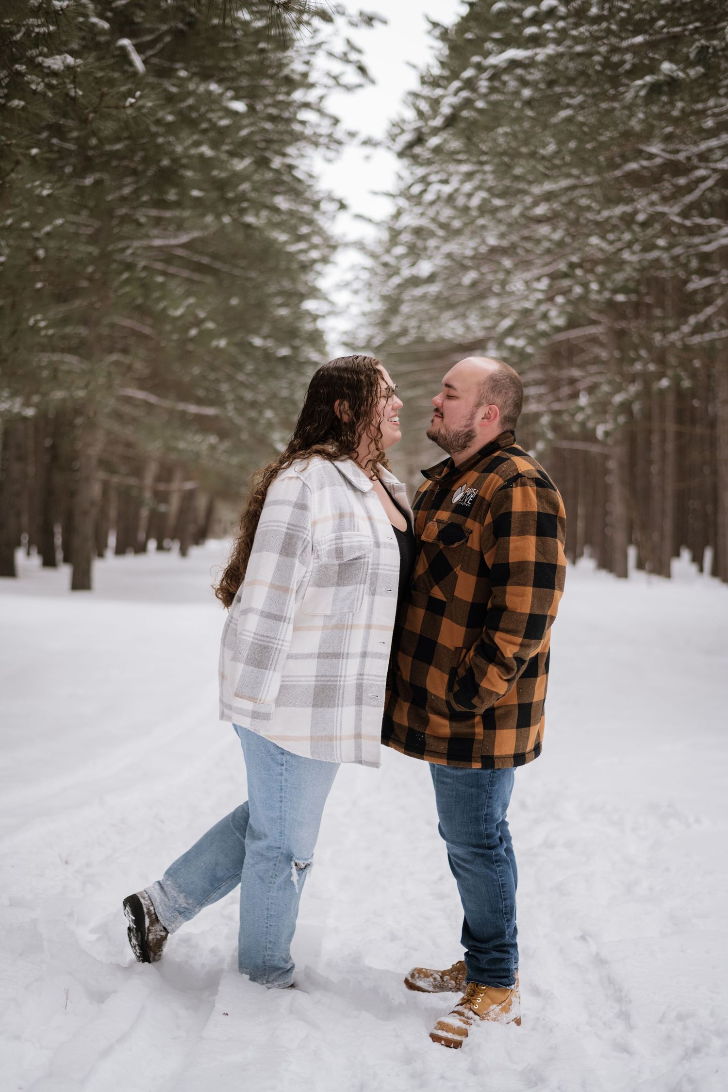 Couple standing face to face on a snowy forest path in plaid jackets, sharing a romantic winter moment.