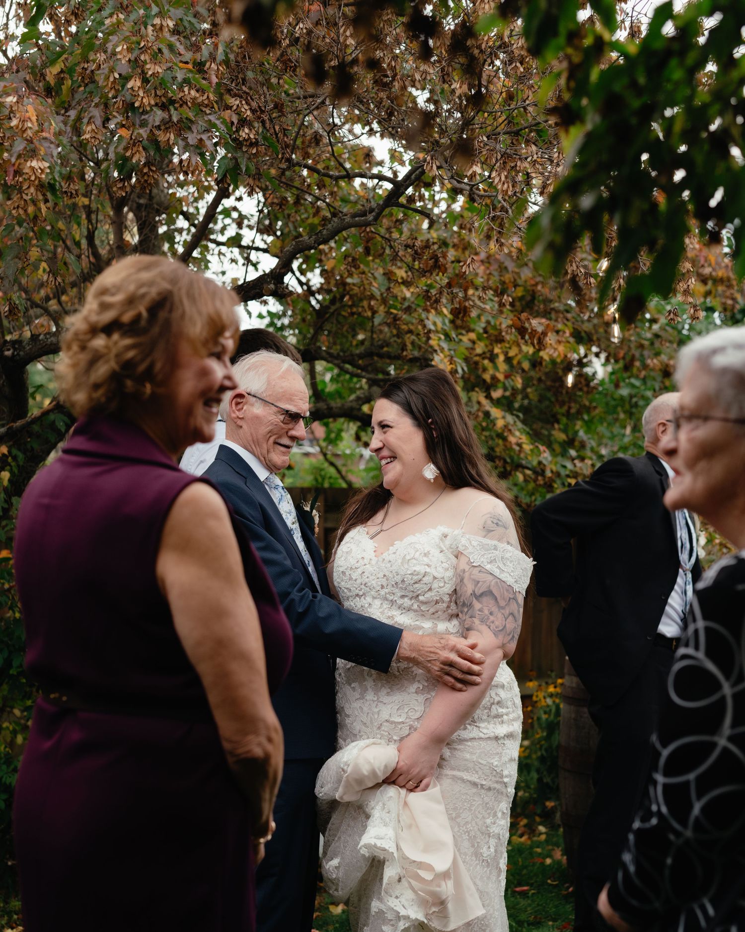 Bride in lace gown laughing with family outdoors at autumn garden wedding ceremony.