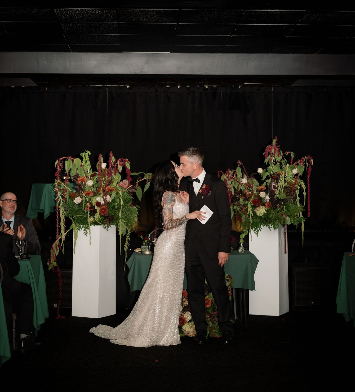 Bride and groom share first kiss at wedding altar adorned with lush burgundy and green floral arrangements on white pedestals.