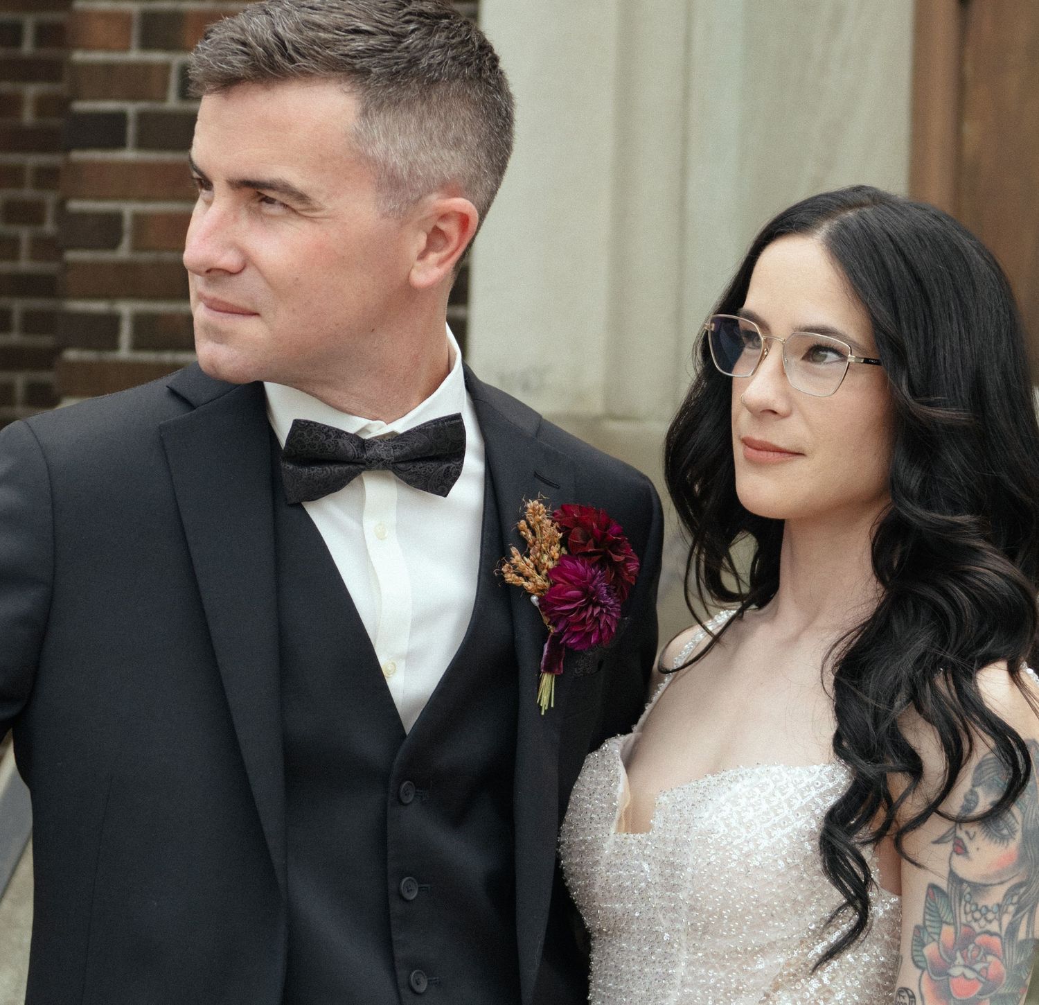 Elegant couple in wedding attire, groom in black tuxedo with floral boutonniere, bride in white gown with tattoos and glasses.