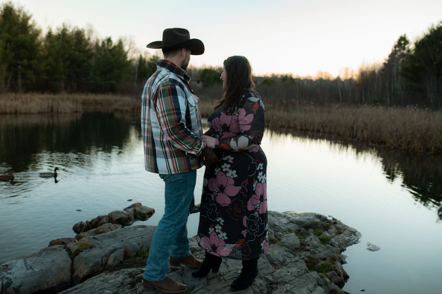 Two people stand at the edge of a tranquil lake during sunset, silhouetted against the calm water and trees.