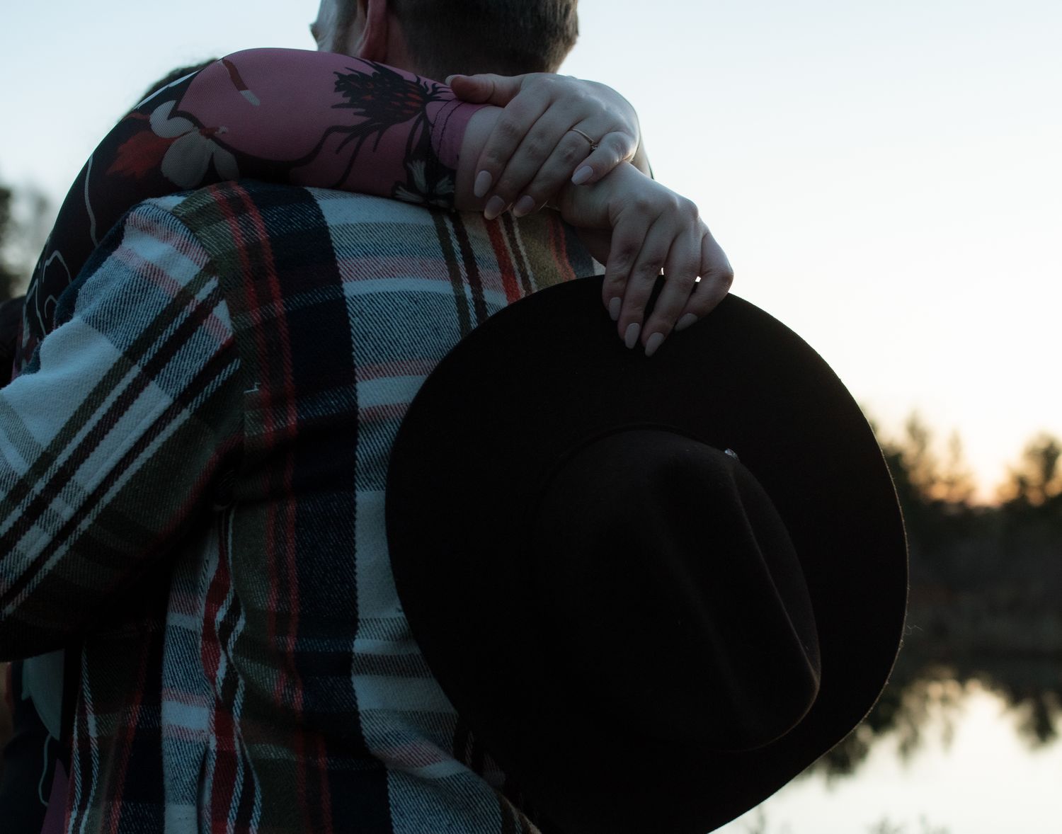 Someone in a plaid shirt holds a black fedora hat against a sunset backdrop near water.