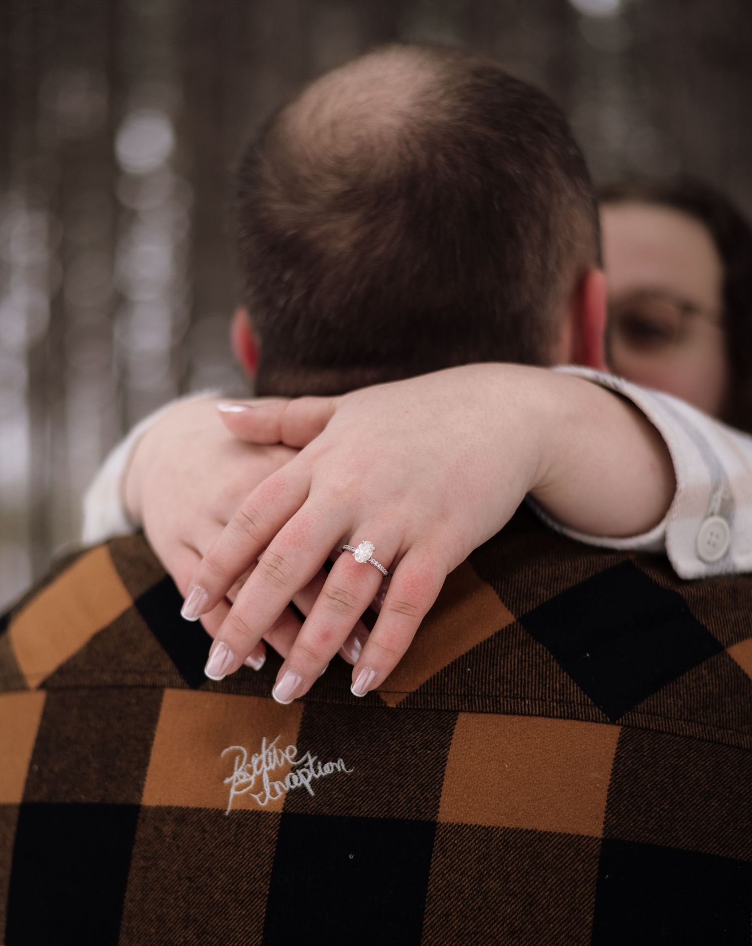 Couple embracing outdoors, woman showing off diamond engagement ring on man's plaid jacket shoulder.