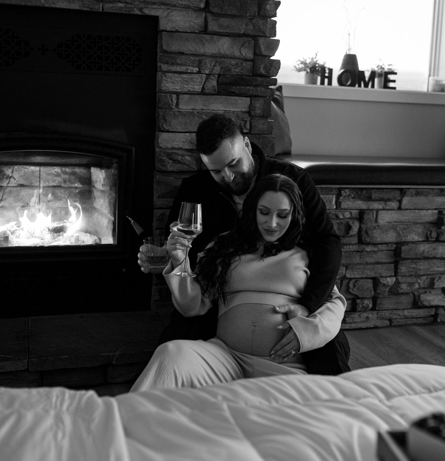 Black and white photo of expecting couple sitting by a glowing fireplace, man holding a wine glass.