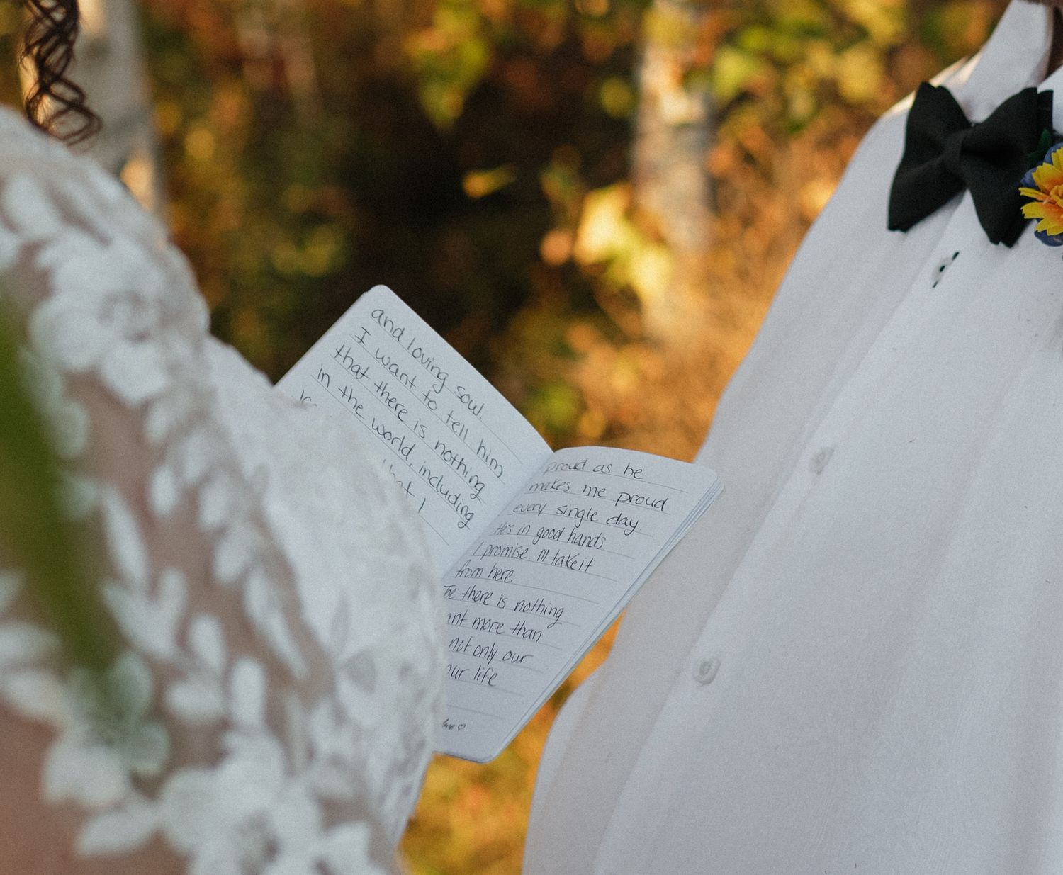 Bride reading handwritten wedding vows from a small notebook, wearing lace dress outdoors in autumn setting.