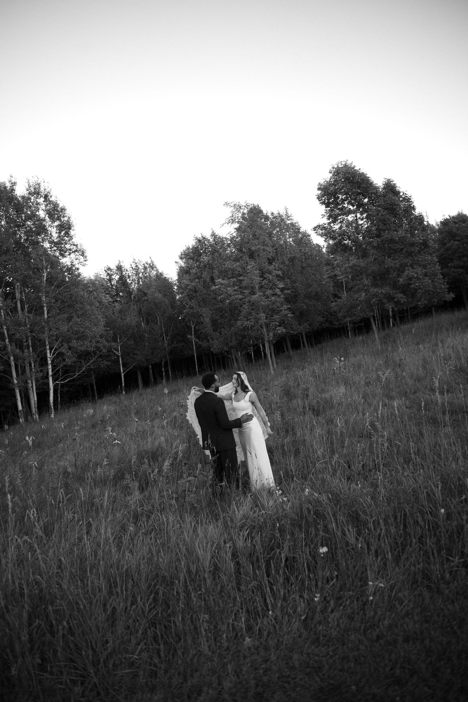 Black and white photo of bride and groom embracing in a meadow surrounded by tall grass and trees.
