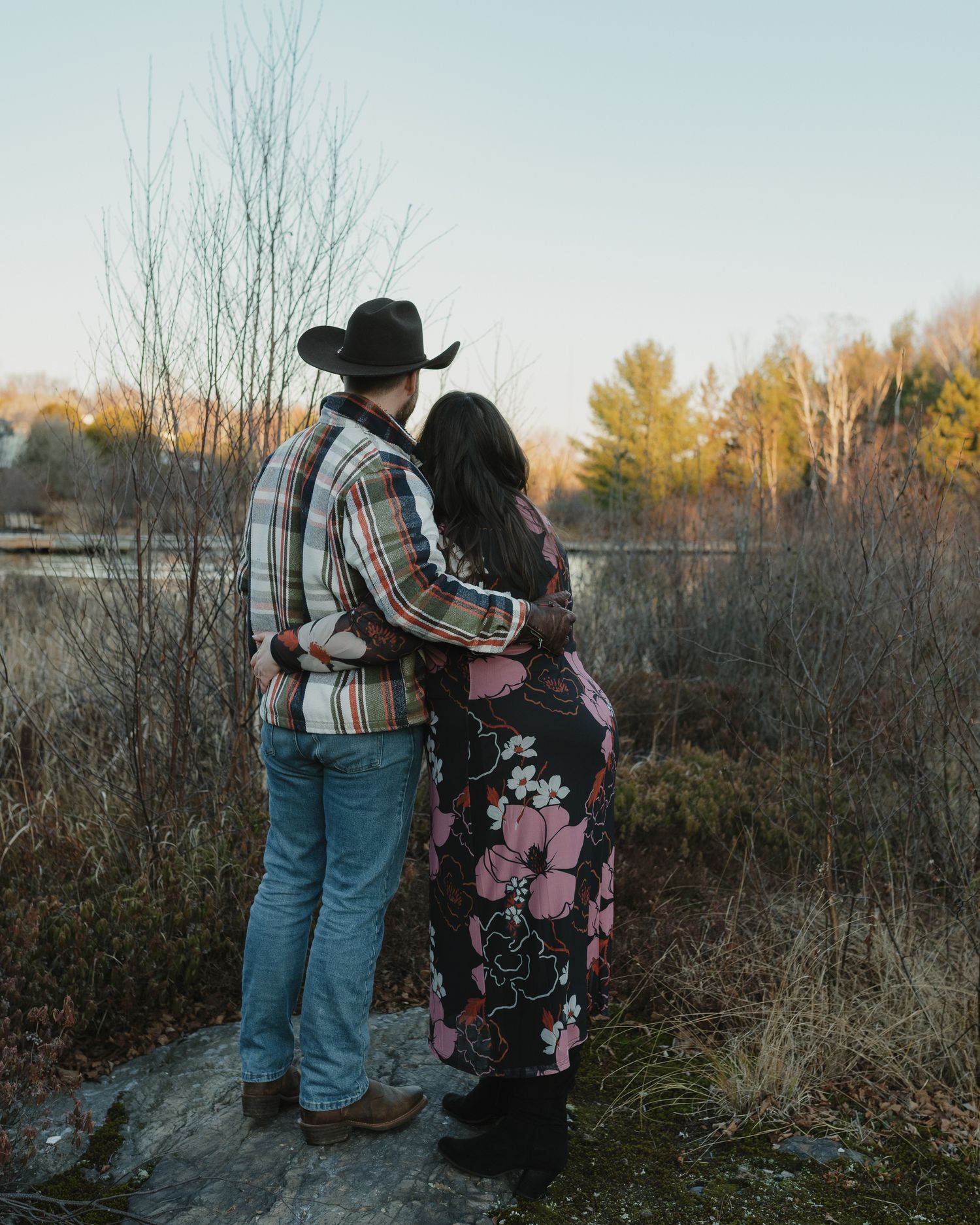 A couple in western attire embraces while looking out over a scenic autumn landscape at sunset.