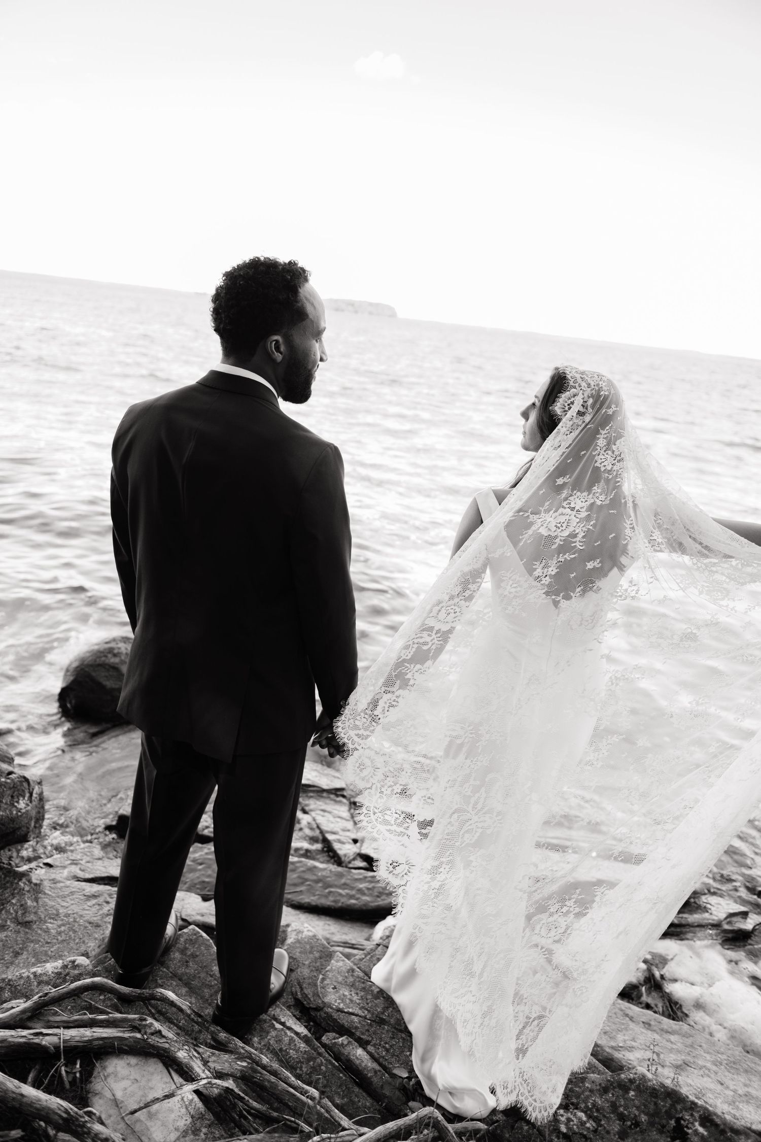 Black and white photo of a bride and groom holding hands on rocky shore, her lace veil flowing in the breeze.