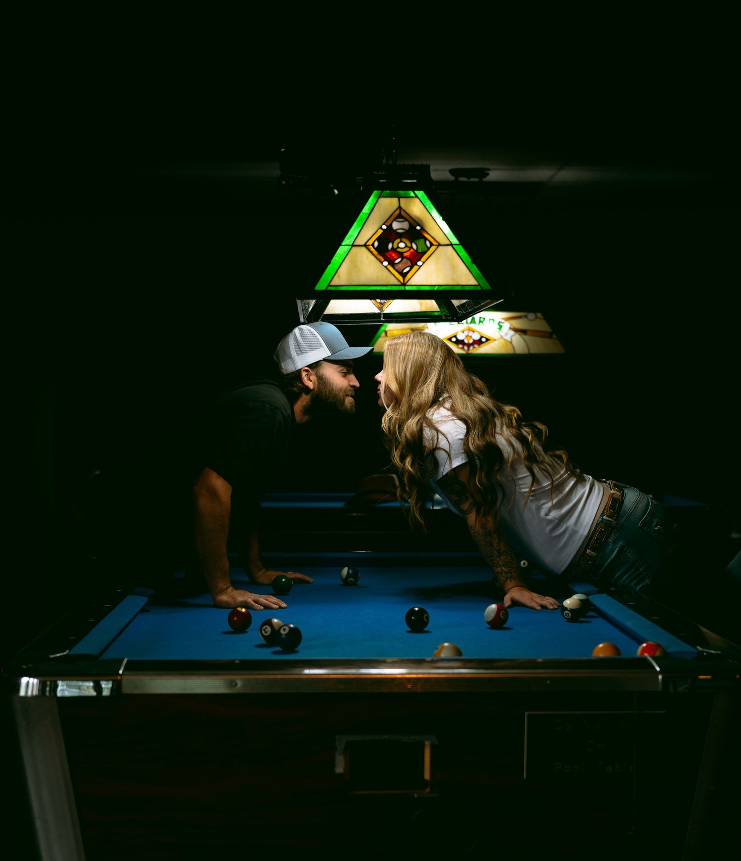 Couple sharing a romantic kiss leaning over a blue pool table under a stained glass billiards lamp in a dark room.