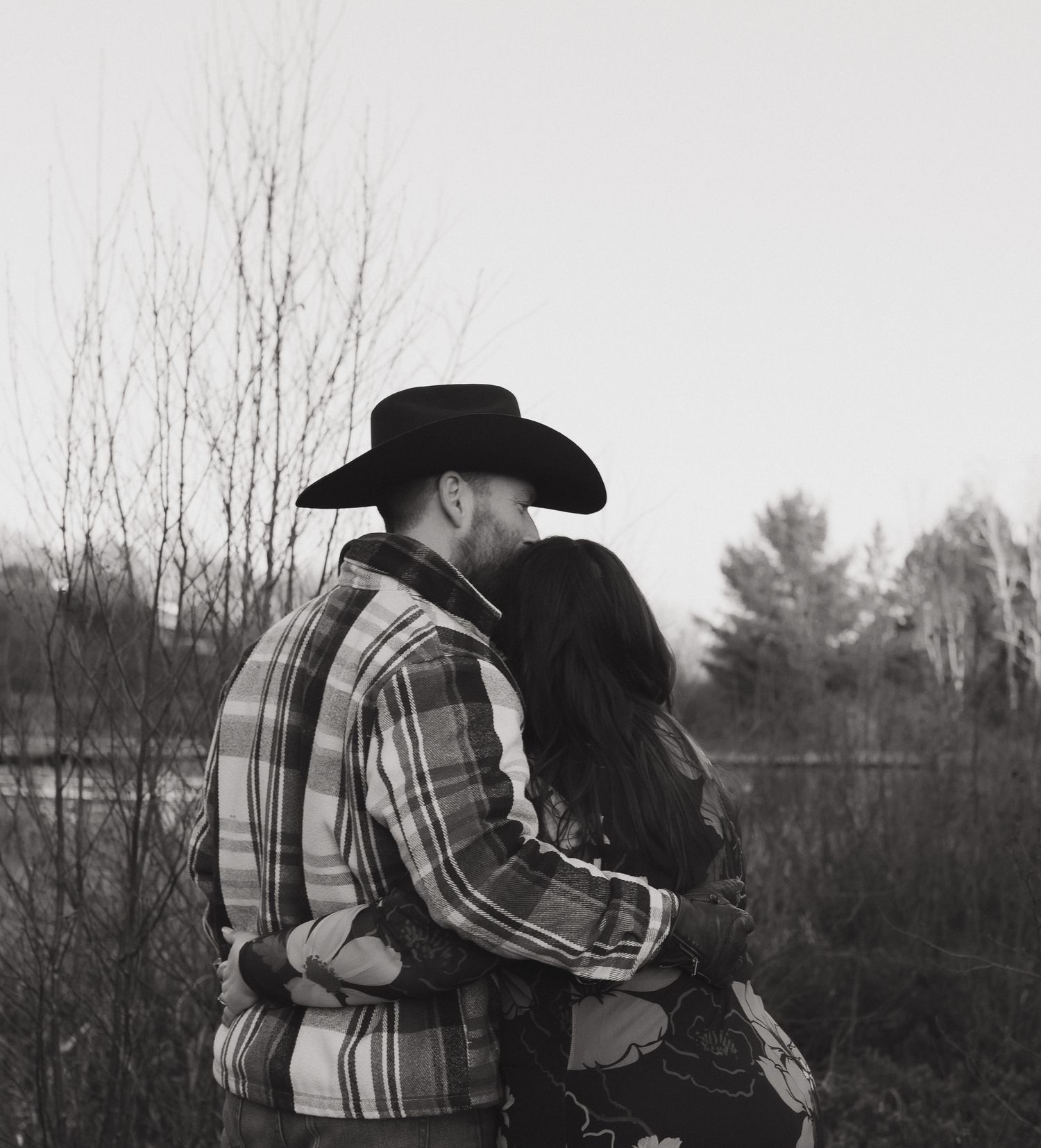A couple embraces in a black and white photo, with one wearing a cowboy hat and plaid jacket against a rural backdrop.