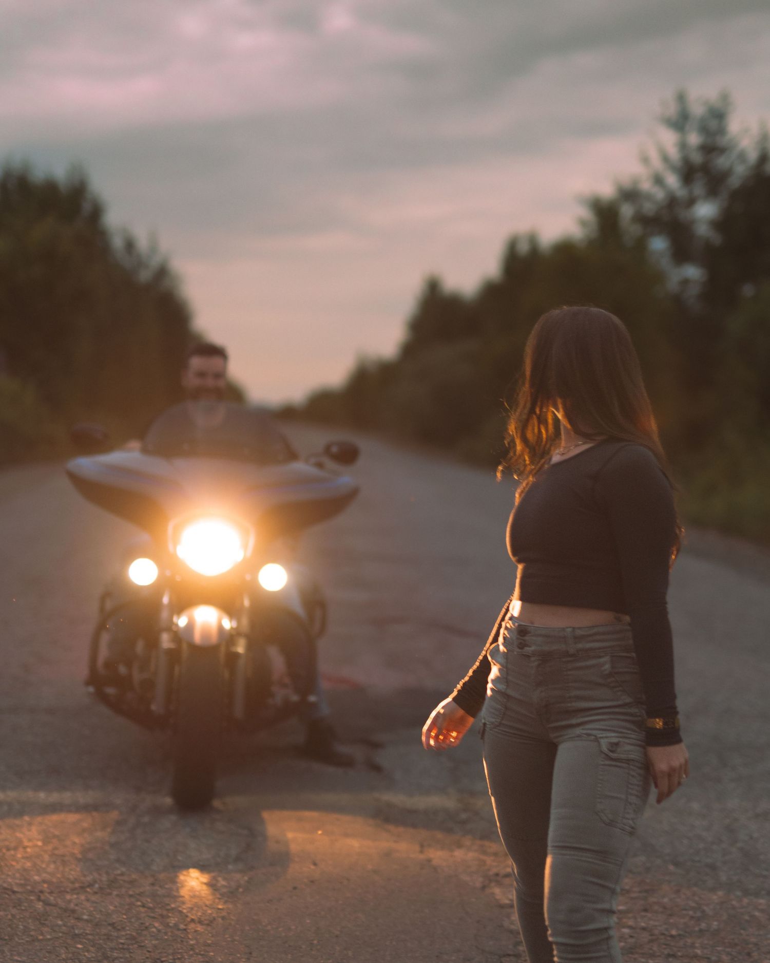 A silhouette of someone watching a motorcycle with headlights on a dusky road.