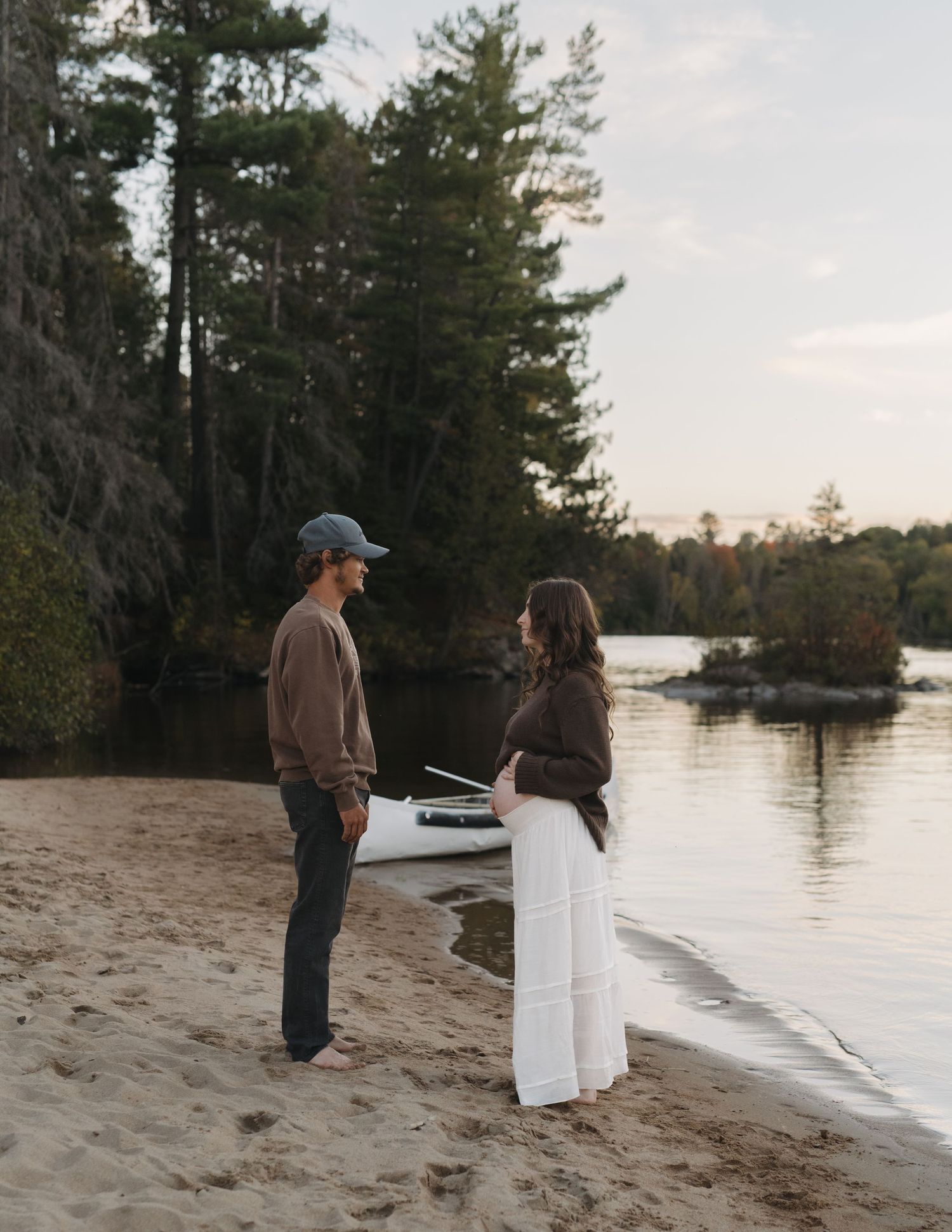 Couple standing on sandy beach at sunset near calm lake waters.