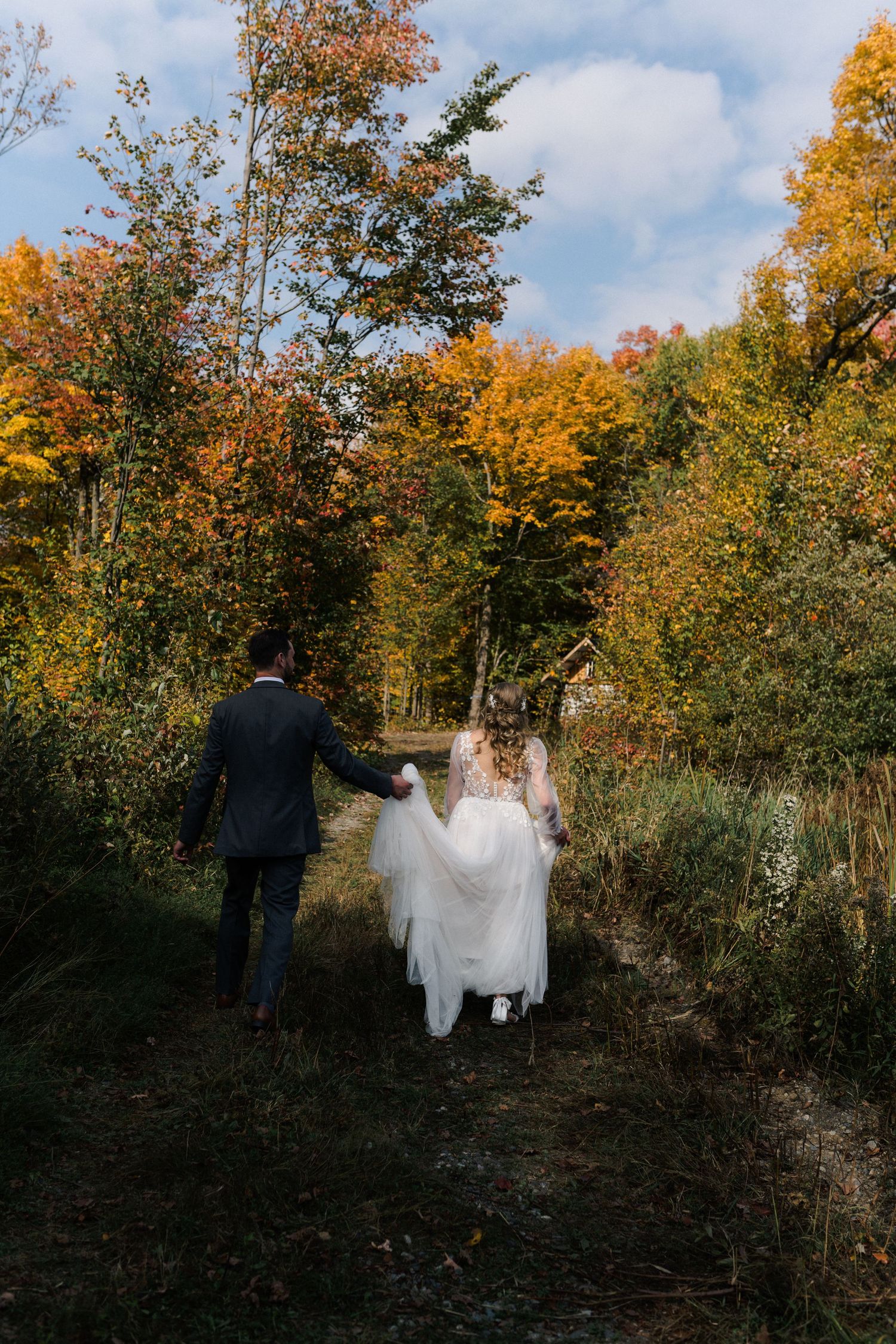 Couple in wedding attire walking through autumn forest with golden foliage and blue sky.