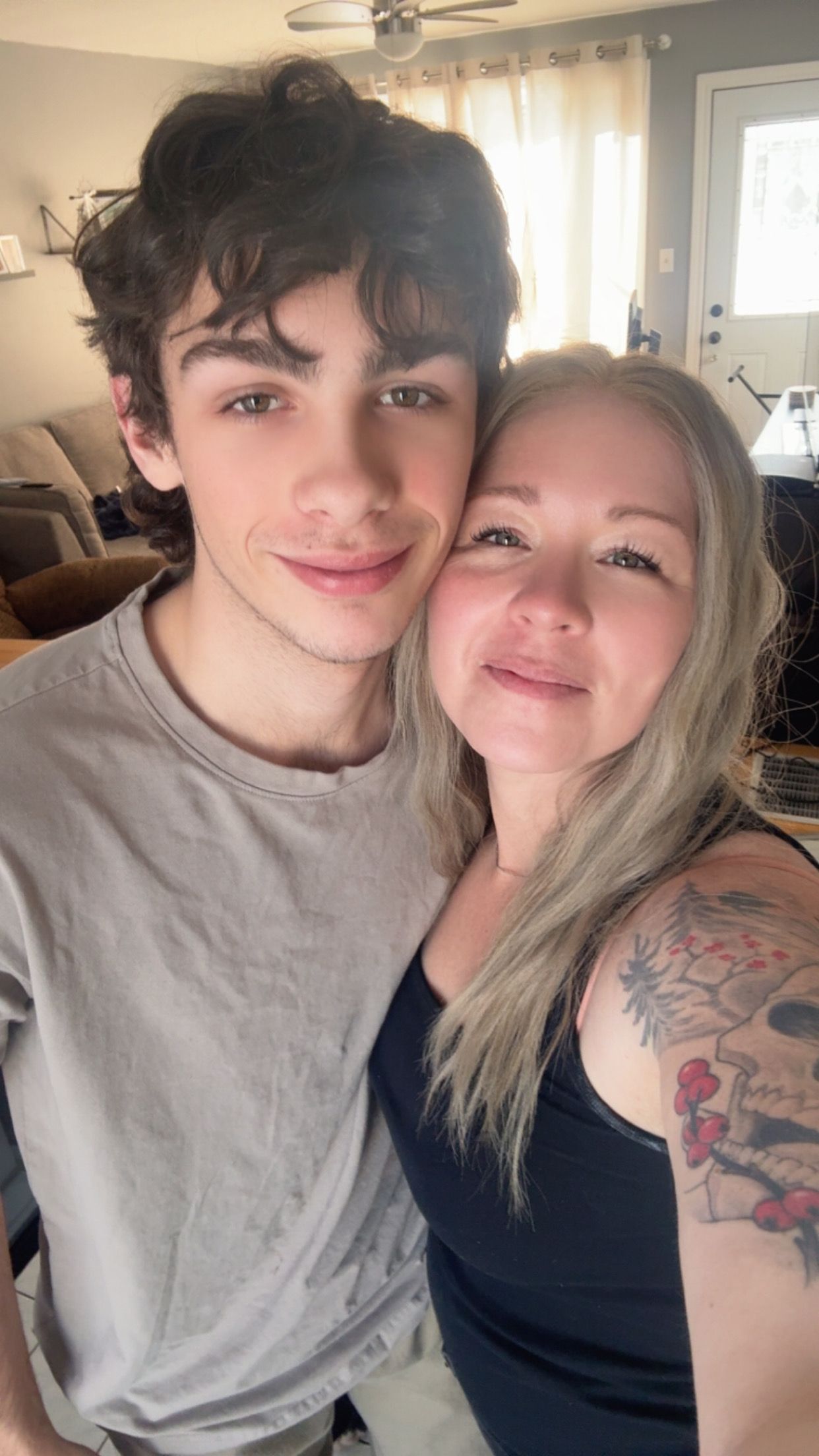 Young man and blonde woman with floral tattoo smile together in a cozy home selfie.