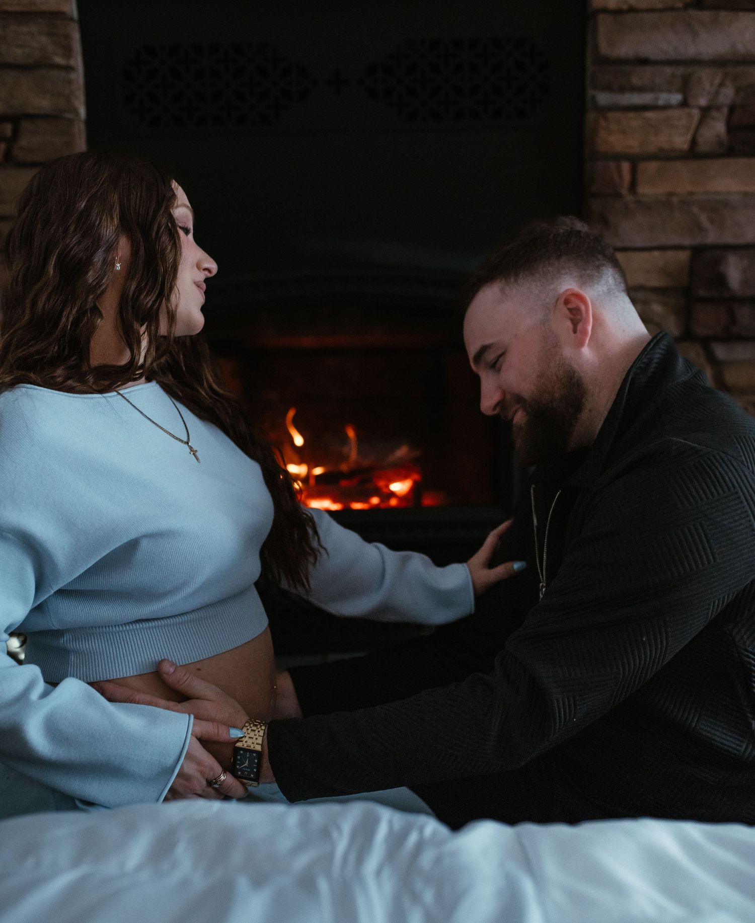 Man tenderly touching pregnant belly while couple sits by glowing fireplace during maternity shoot.