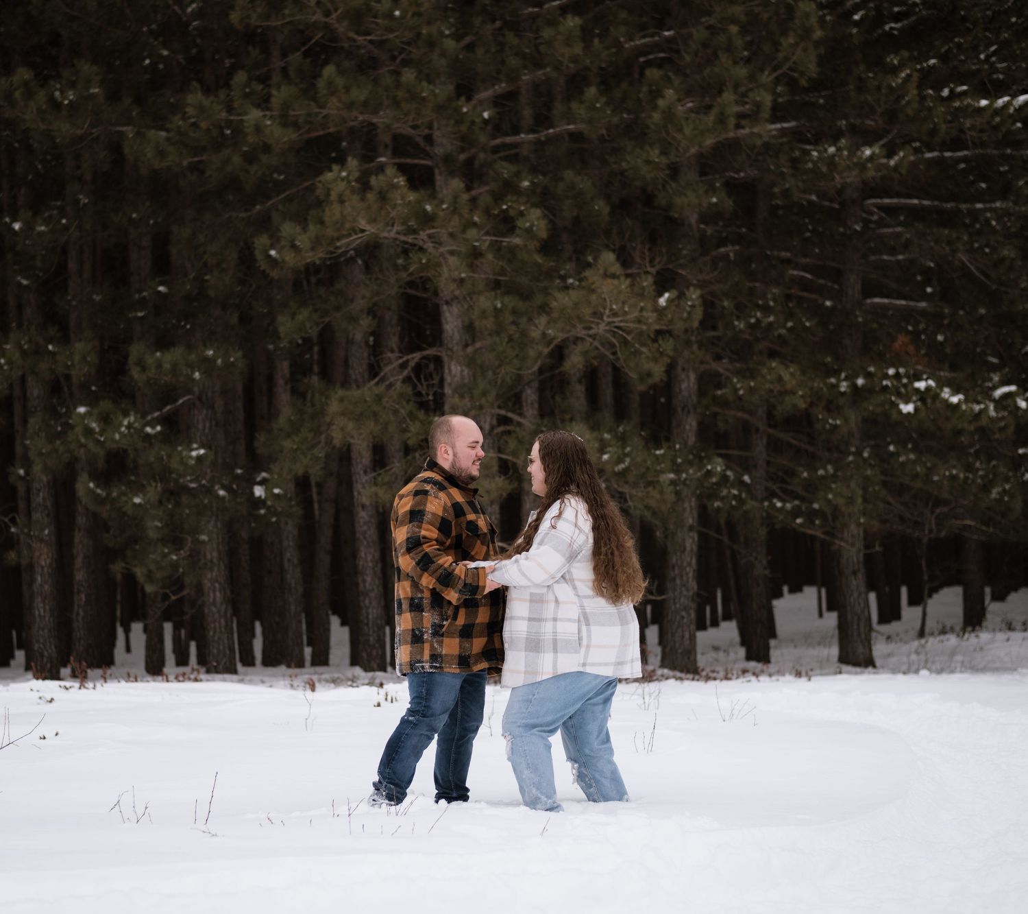 Couple holding hands in snowy winter forest with tall pine trees, wearing plaid shirts and jeans.