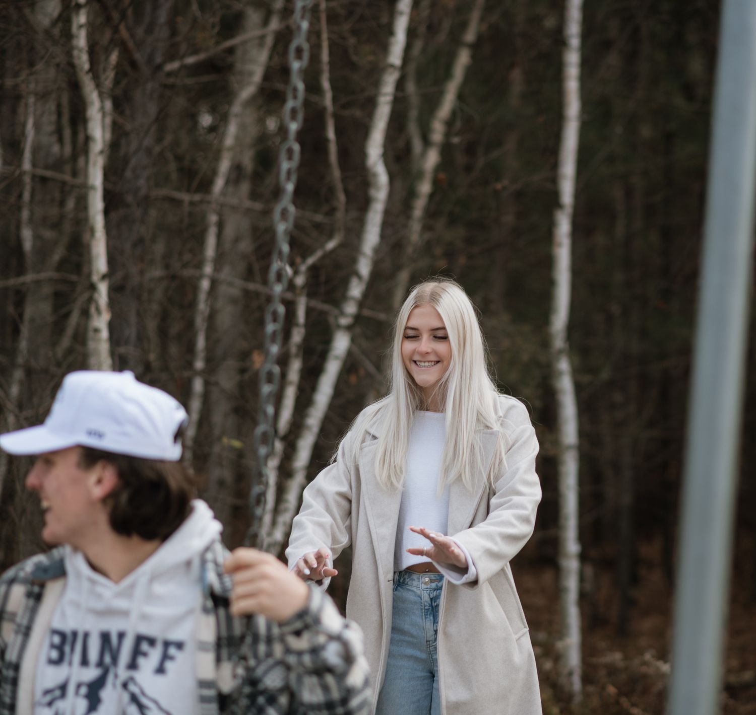 Smiling blonde woman in a beige coat reaching out to companion in a forest setting.