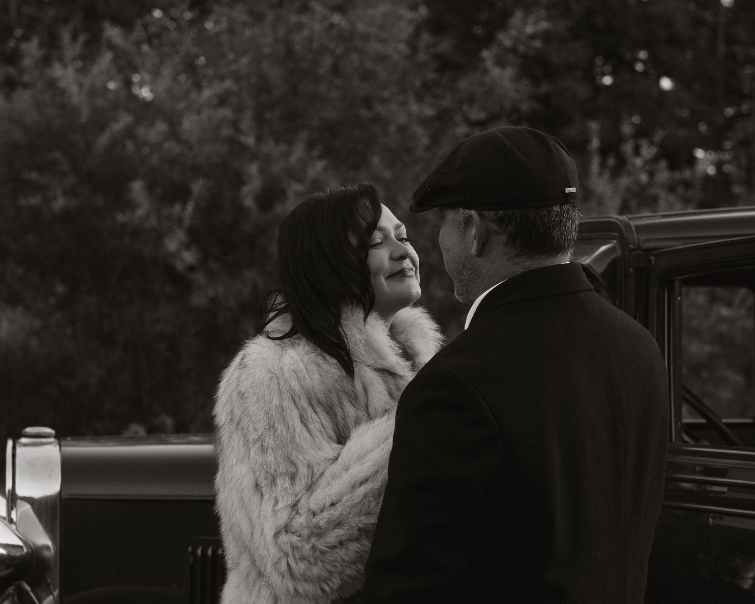 Black and white photo of a woman in fur coat and man in cap sharing an intimate moment near a vintage car.