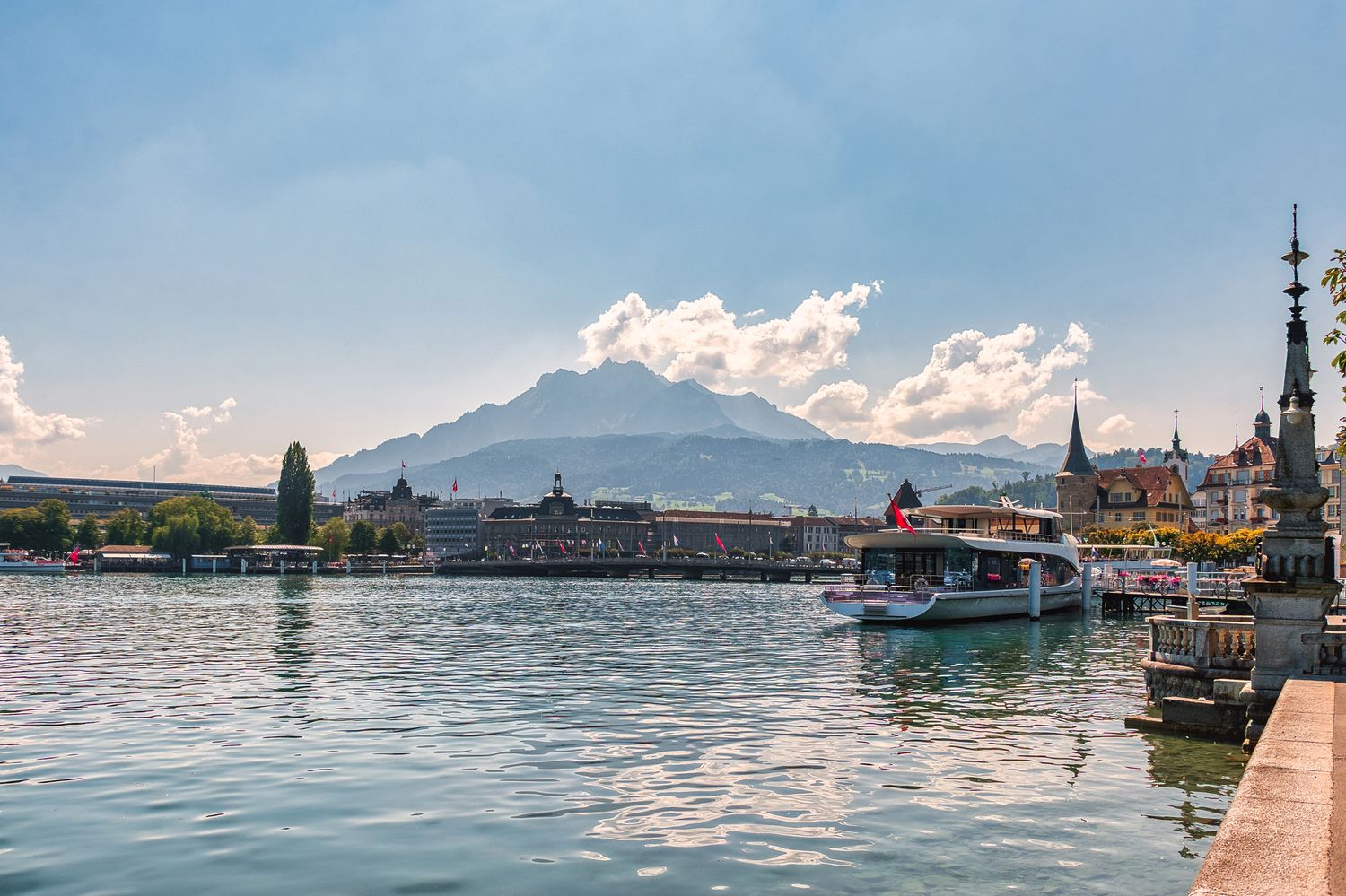 Scenic view of Lucerne waterfront with boats, historic buildings, and Mount Pilatus under a partly cloudy sky.