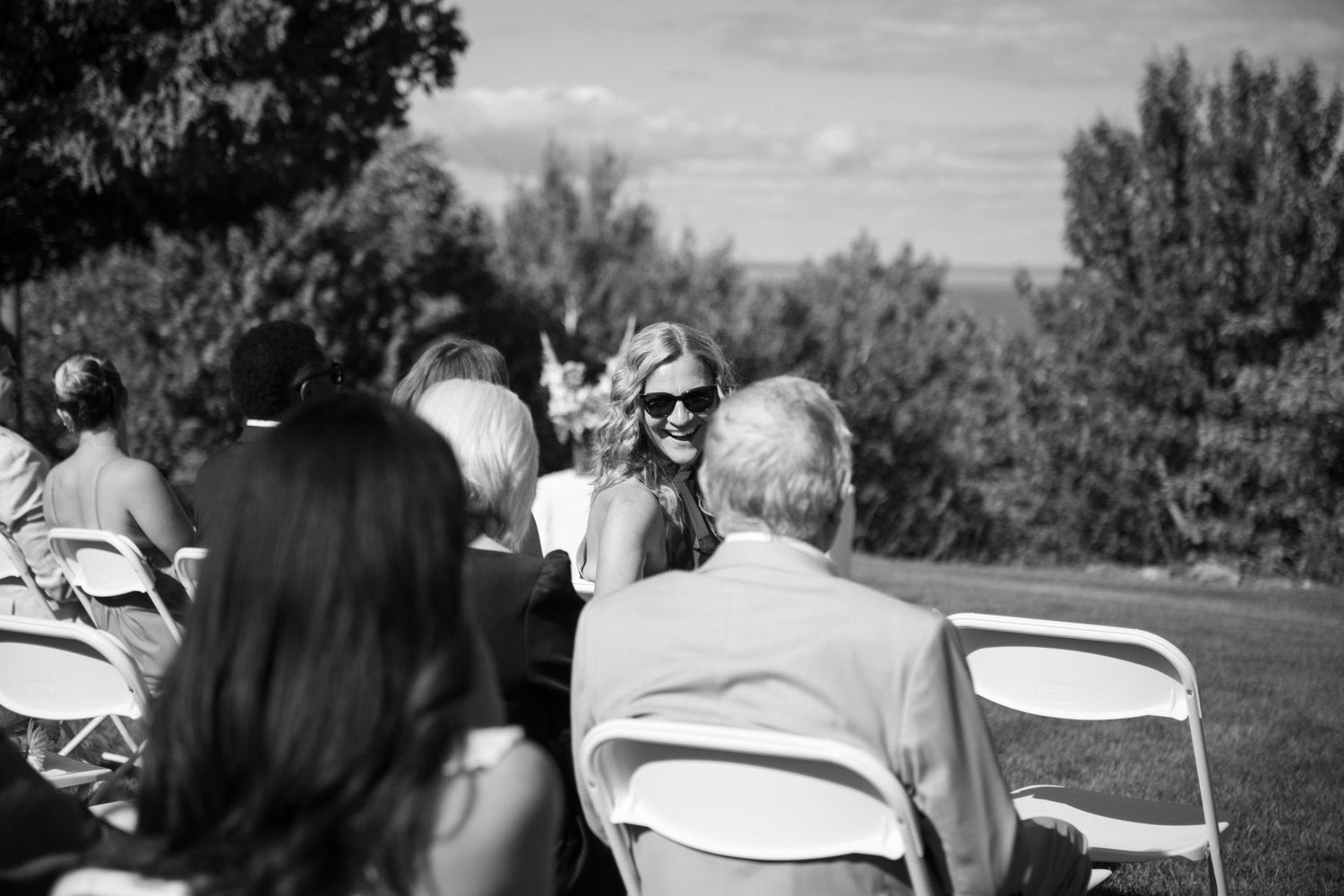 Black and white outdoor wedding ceremony guests seated on white folding chairs in a sunny garden setting.