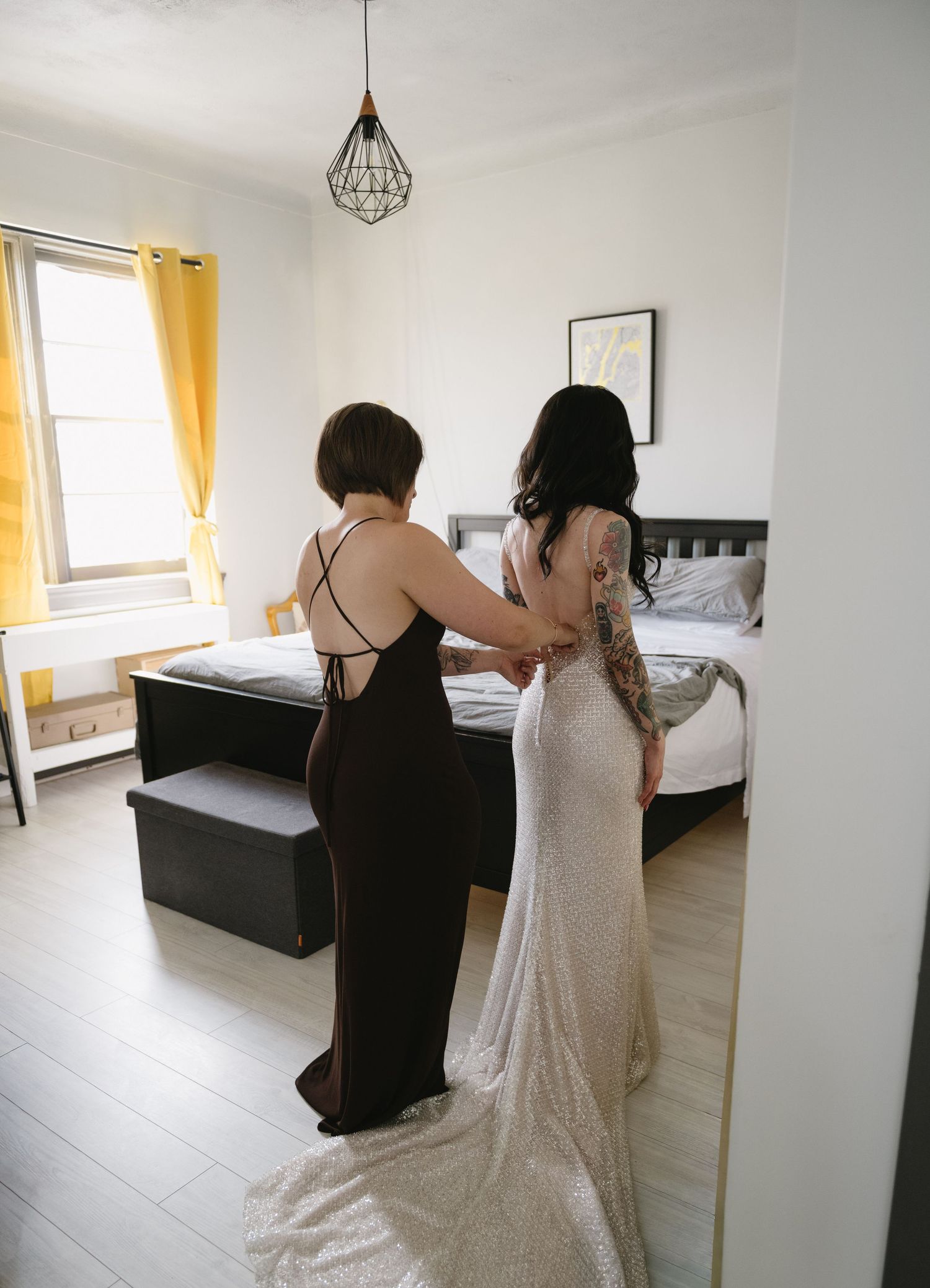 A bride in a sequined gown gets help from a bridesmaid in a black dress in a bright bedroom before the wedding.