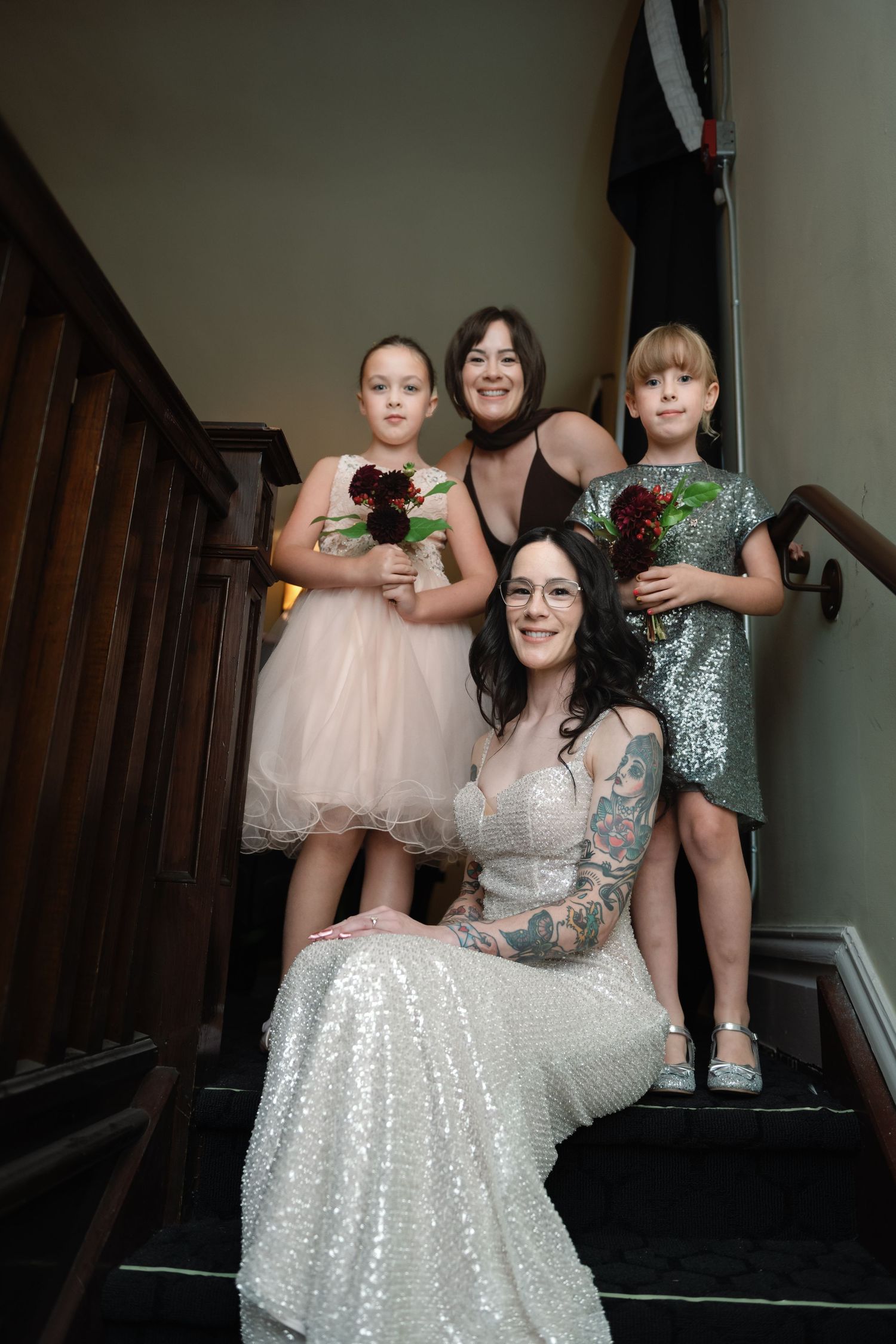 Bride in sequin gown sits on staircase with tattooed arms, flanked by two flower girls and a bridesmaid holding bouquets.