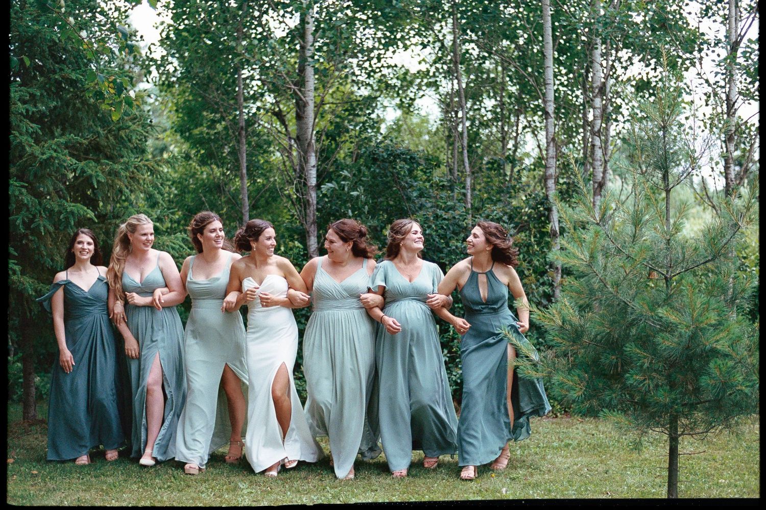 Bridesmaids in sage green dresses walking together outdoors near trees with bride in white gown at a wedding.