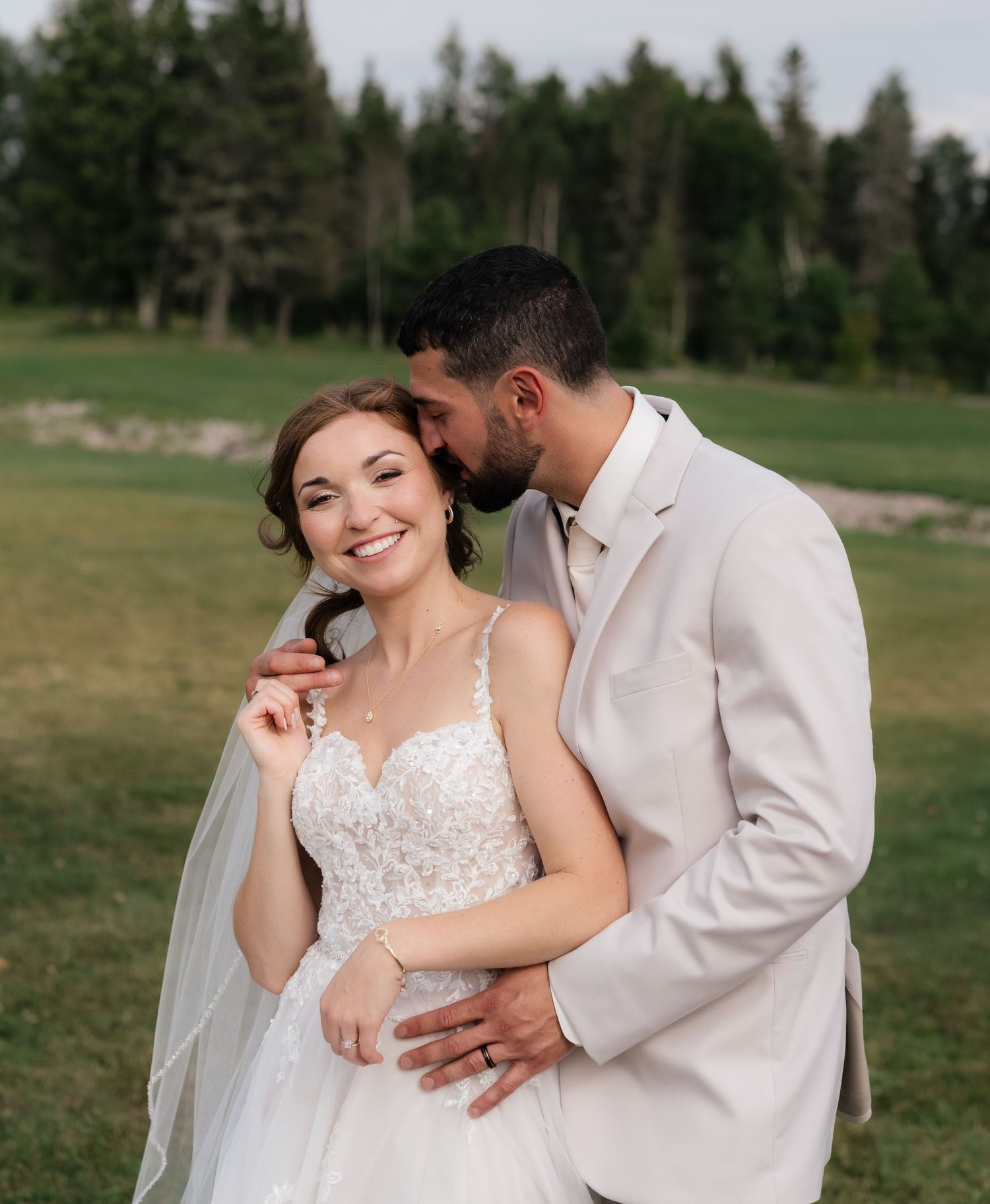 Couple in wedding attire share intimate moment on grassy field with forest backdrop.