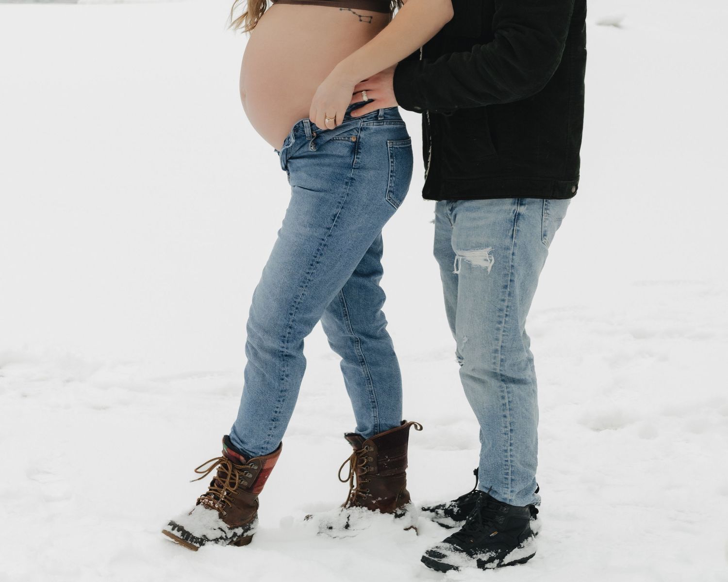 A tender maternity moment captured in the snow with blue jeans and winter boots.