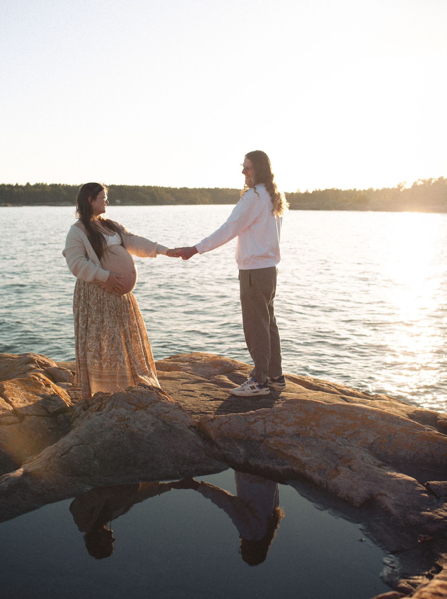 Couple sitting on rocky shoreline at sunset overlooking a serene lake.