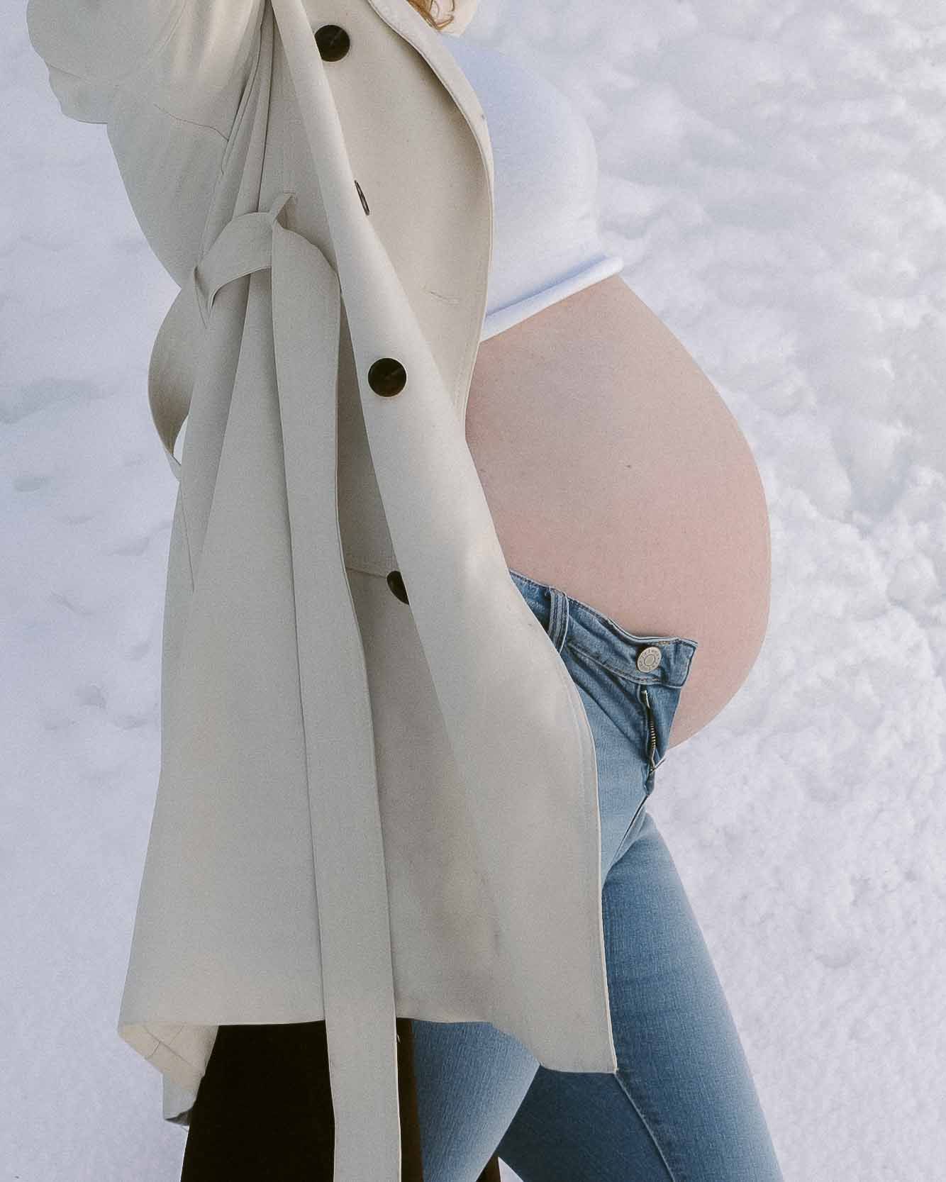 A person in a light coat and jeans stands against a snowy background.