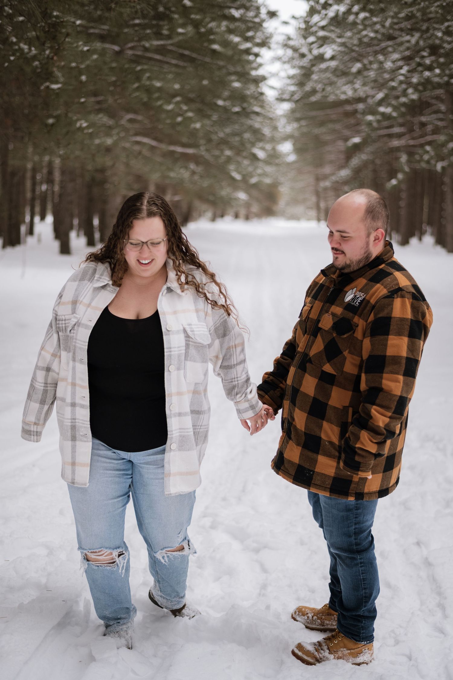 Couple holding hands walking on a snowy forest path in plaid flannel shirts and jeans.