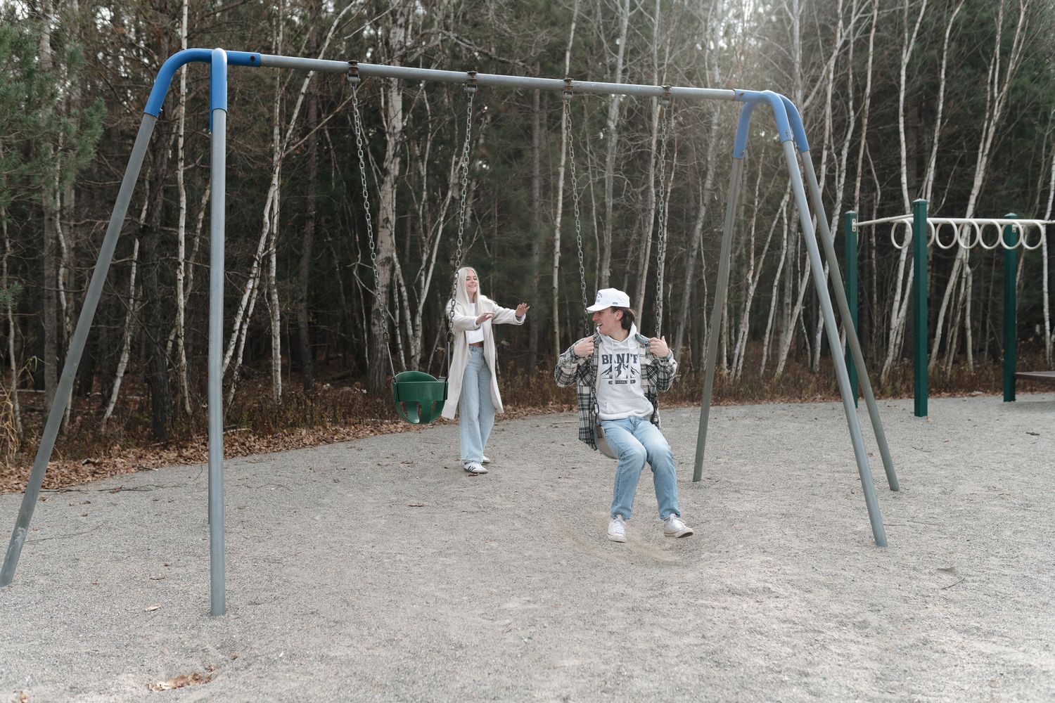 Two children playing on metal swings at a playground surrounded by tall trees in autumn.