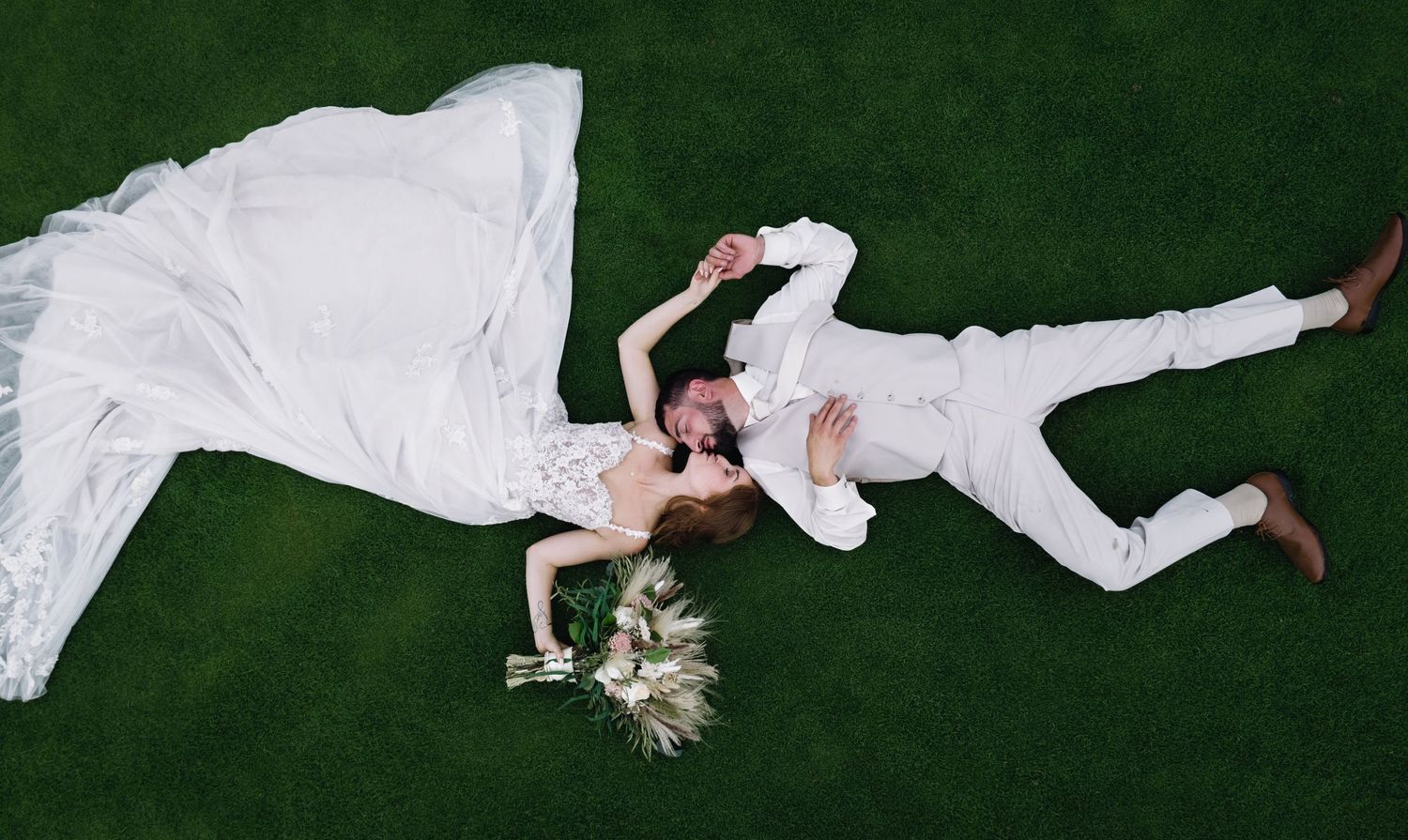 Aerial view of a bride and groom lying on green grass, holding hands, bride in white gown with bouquet.