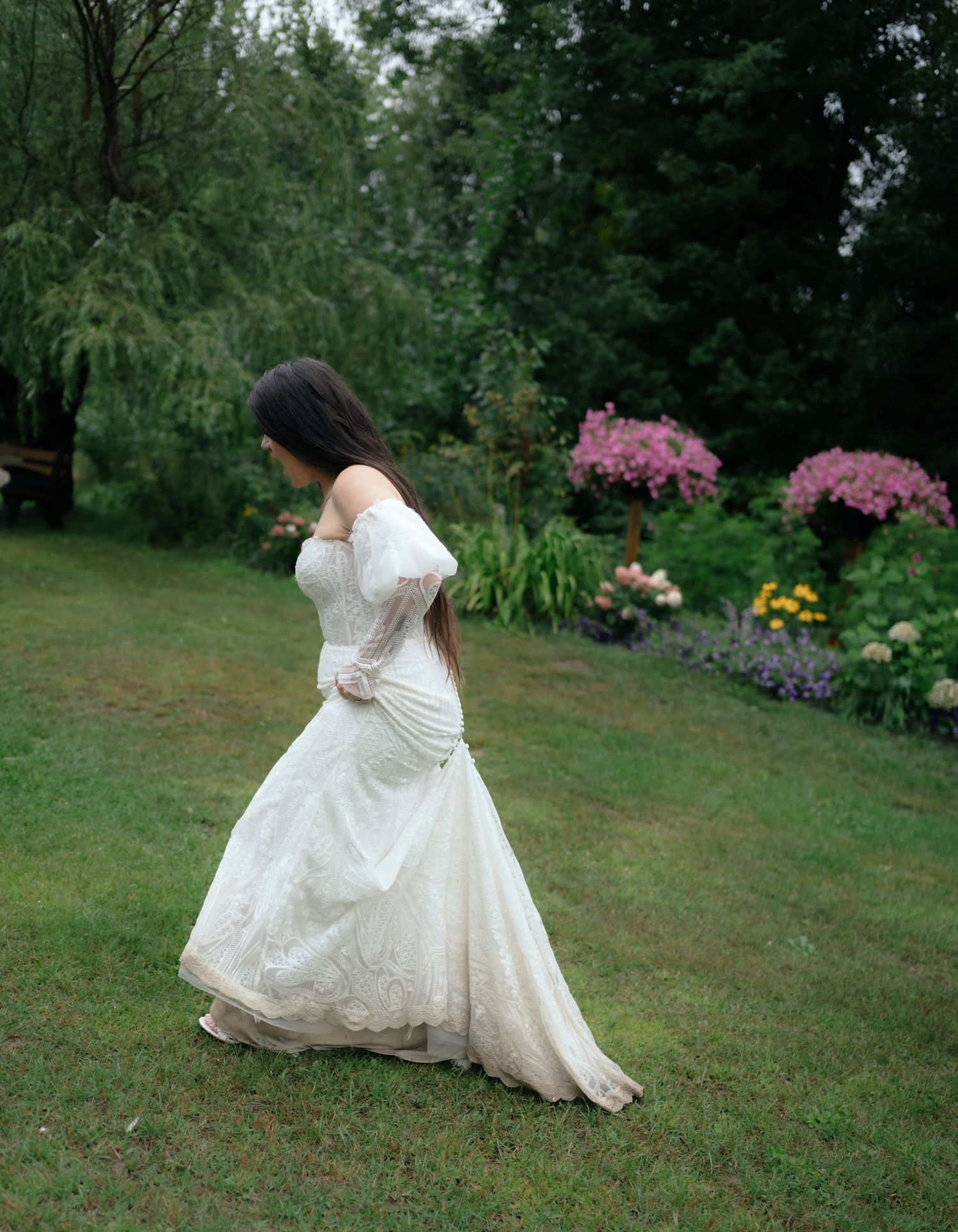 Bride in white lace off-shoulder wedding gown walking gracefully through a lush garden with colorful flowers.