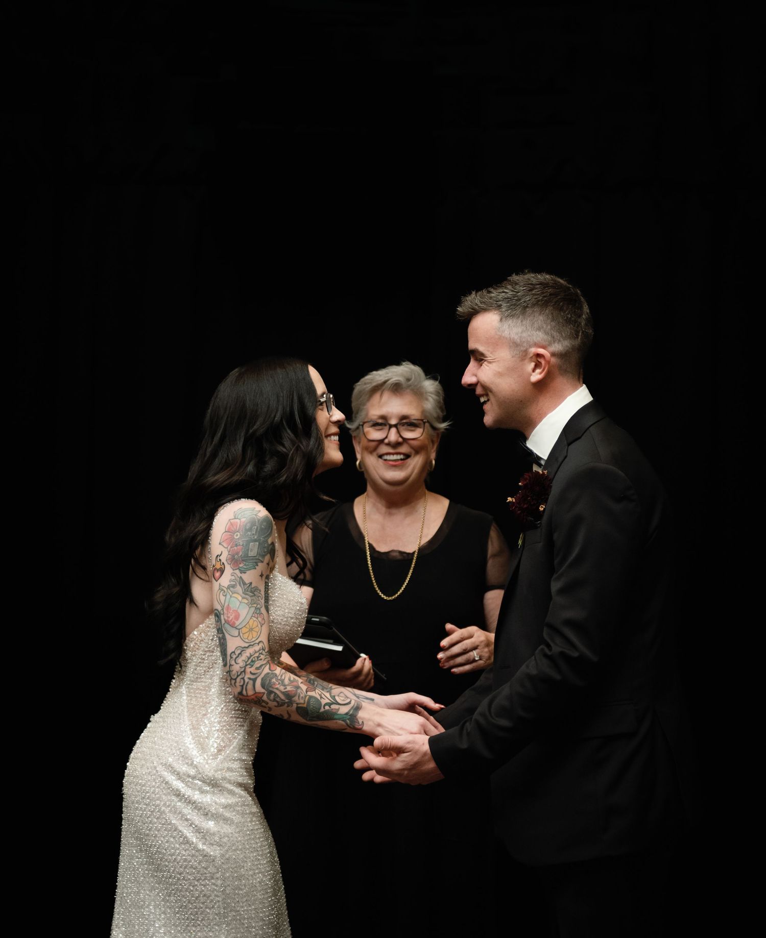 Bride with tattoos and groom holding hands at wedding ceremony with officiant smiling in background.