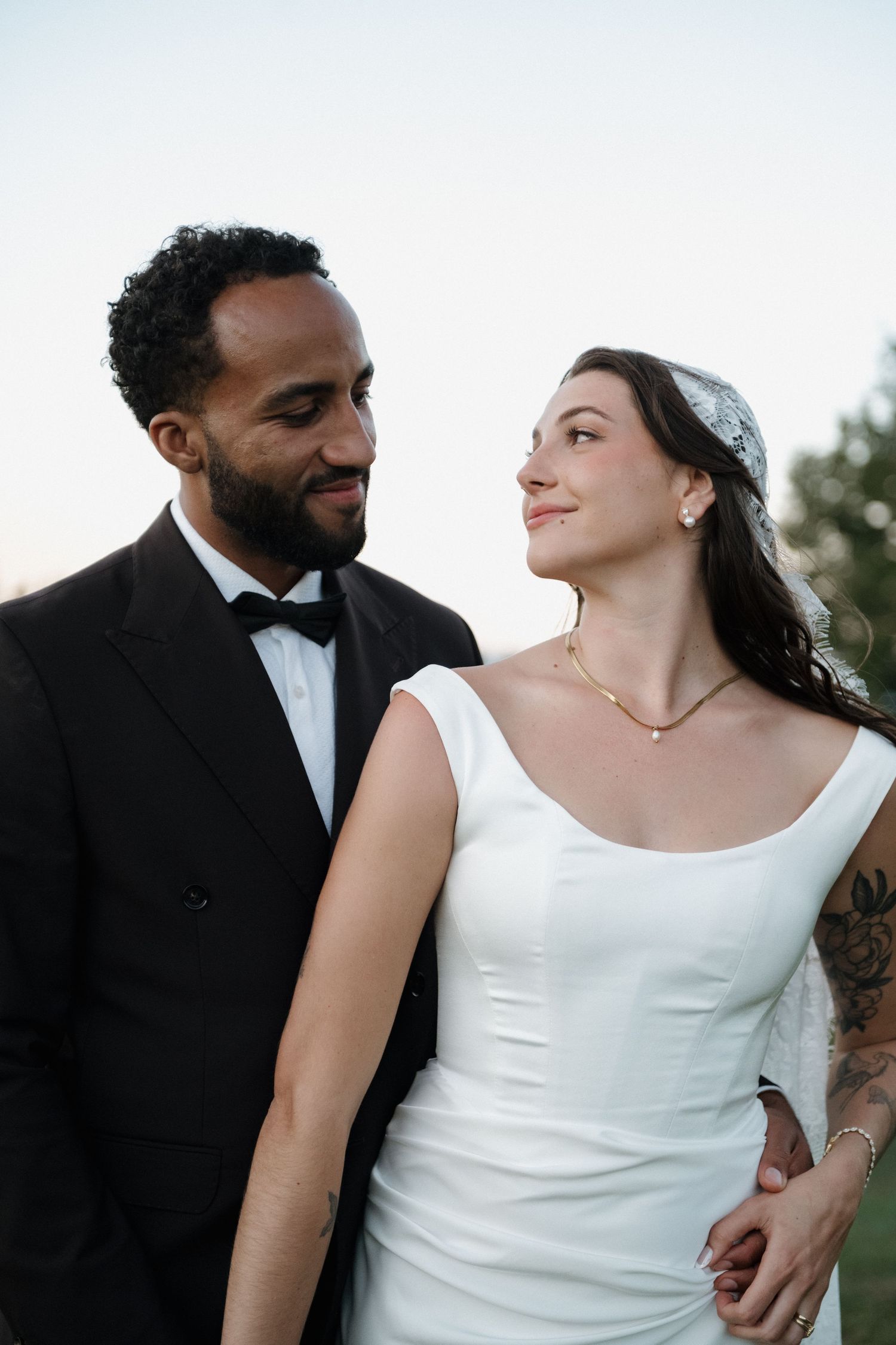 Smiling bride and groom in wedding attire gazing at each other outdoors at dusk.