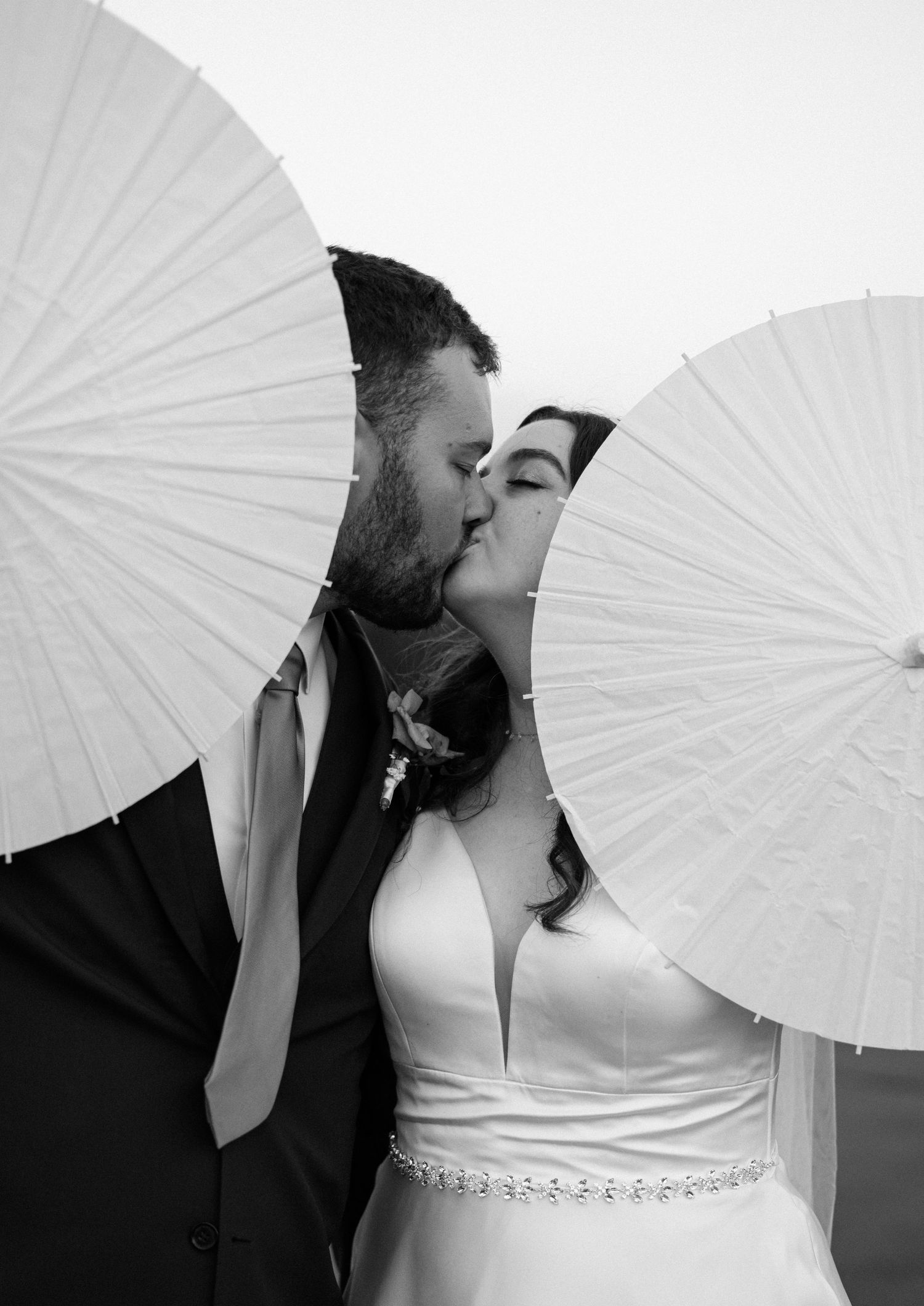 Black and white photo of a bride and groom kissing behind two white parasols on their wedding day.