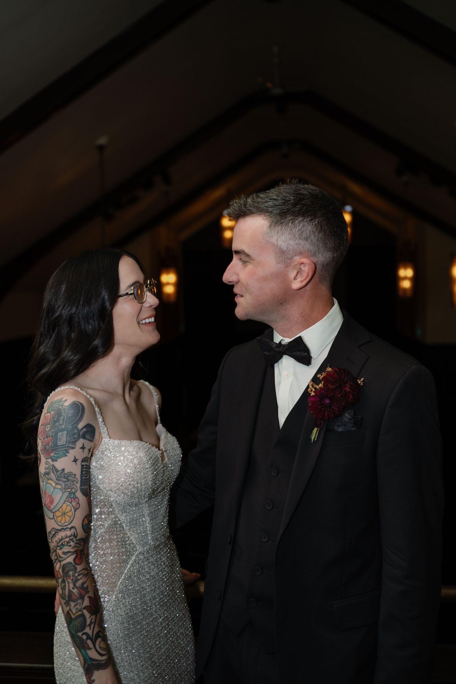 A tattooed bride in a sparkly gown and groom in a black tuxedo share a loving gaze at their wedding ceremony.