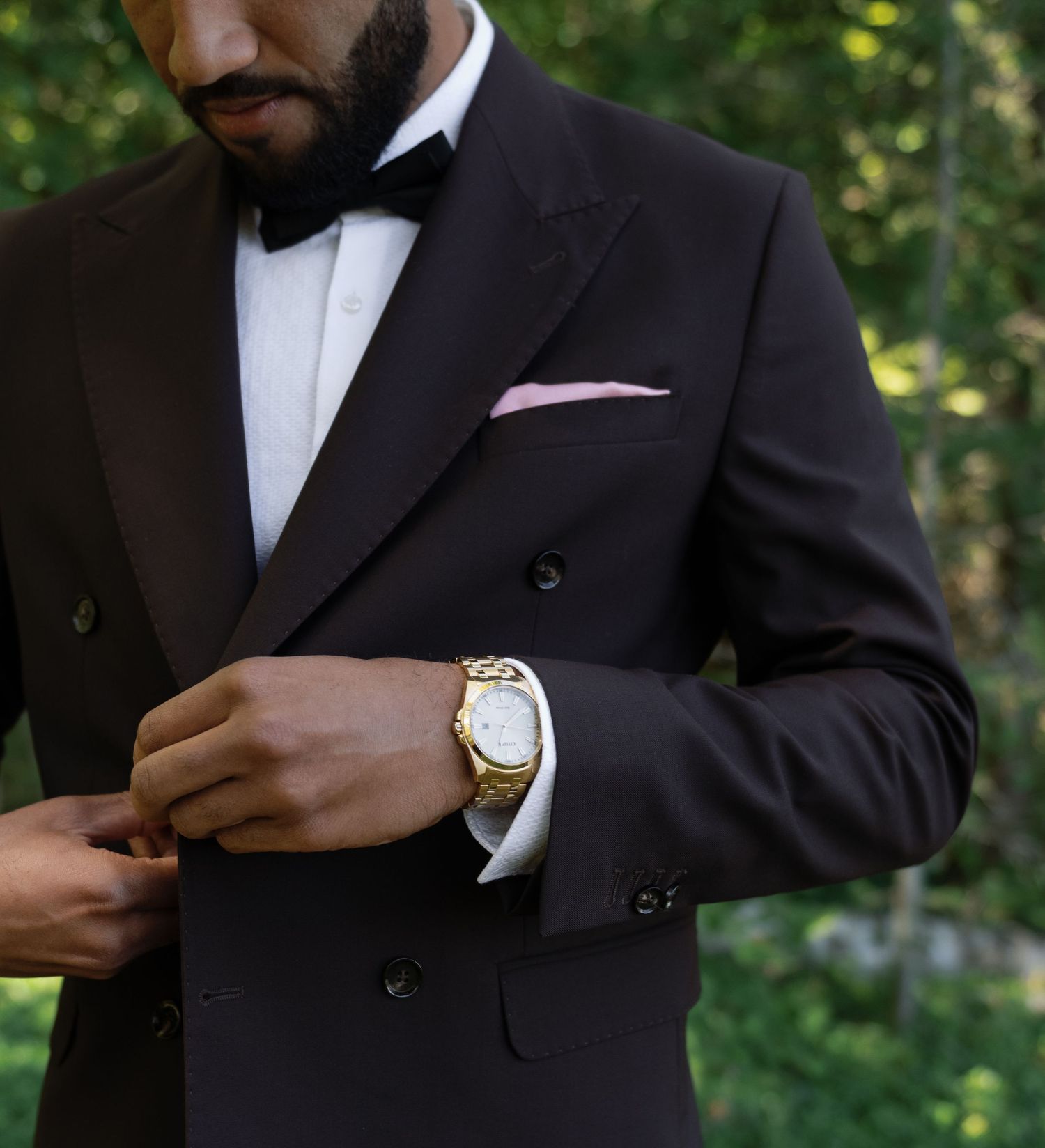 Groom in black tuxedo adjusts gold watch, styled with bow tie and pink pocket square.