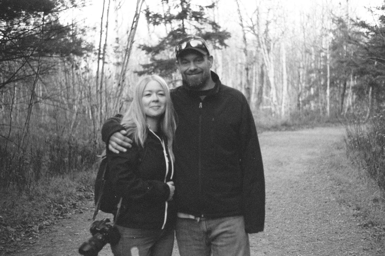 Black and white photo of a smiling couple posing together on a forest path surrounded by bare trees.