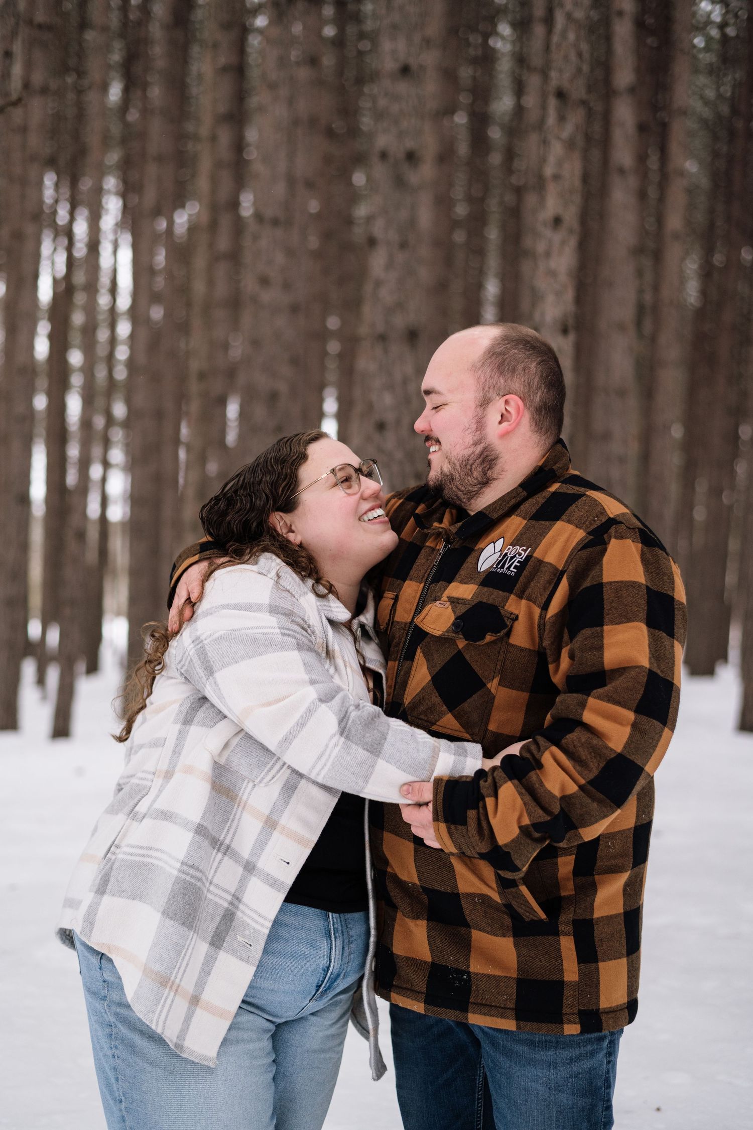 A couple embraces and smiles at each other in a snowy forest, wearing flannel shirts and jeans in winter.