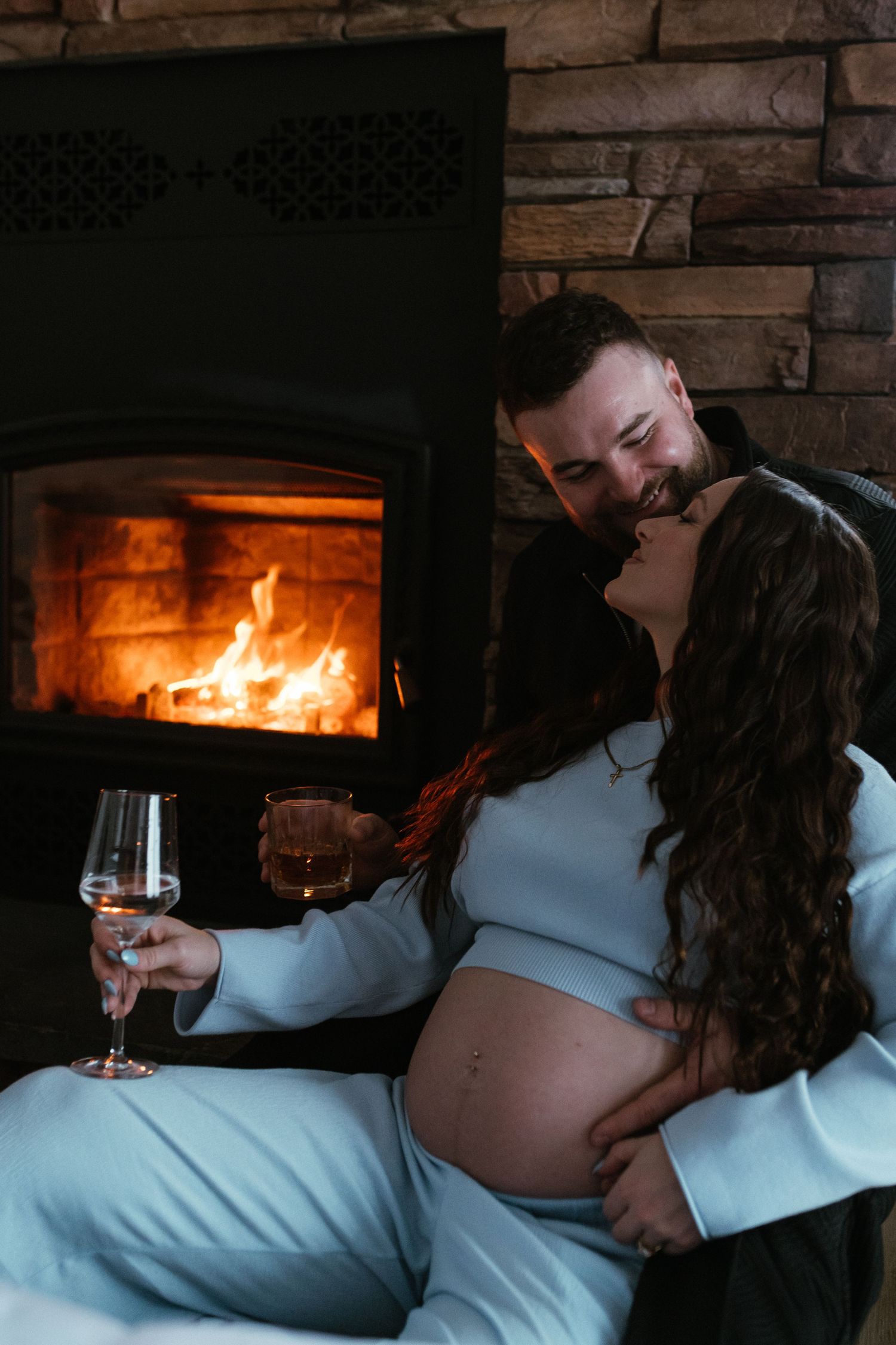Smiling couple sitting by a warm fireplace, pregnant woman in blue outfit holding a wine glass.