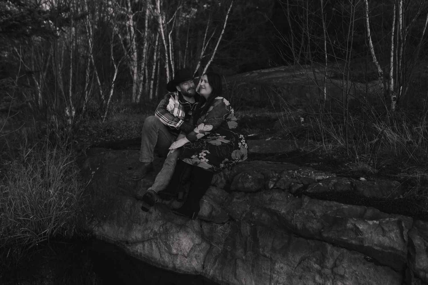A romantic couple sits together on a rocky cliff edge in a moody black and white forest setting at dusk.